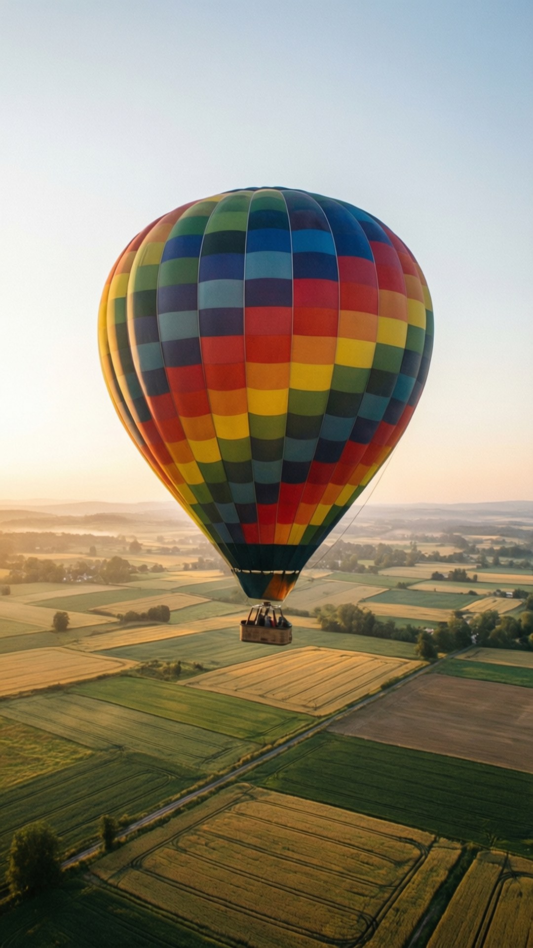 Balloon at Dawn 壁紙 — 一個充滿活力的彩虹熱氣球在日出時分平靜地漂浮在金黃色和綠色的農田拼布之上。