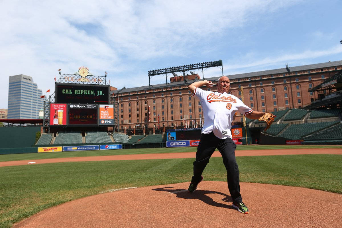 cal ripken jr 桌布 壁紙 — Cal Ripken Jr. 身穿白色金鶯隊球衣，在陽光明媚的球場上投擲棒球，身後是體育場座位和記分牌。
