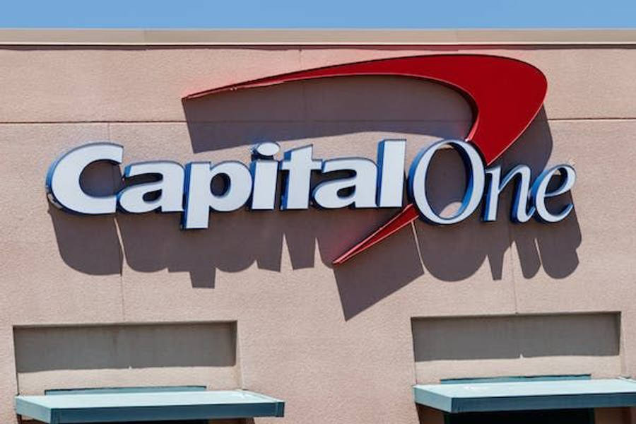 桌布 壁紙 — Capital One 銀行標誌，帶有粗體紅色弧線和白色字體，安裝在米色建築物上，背景是晴朗的藍天。