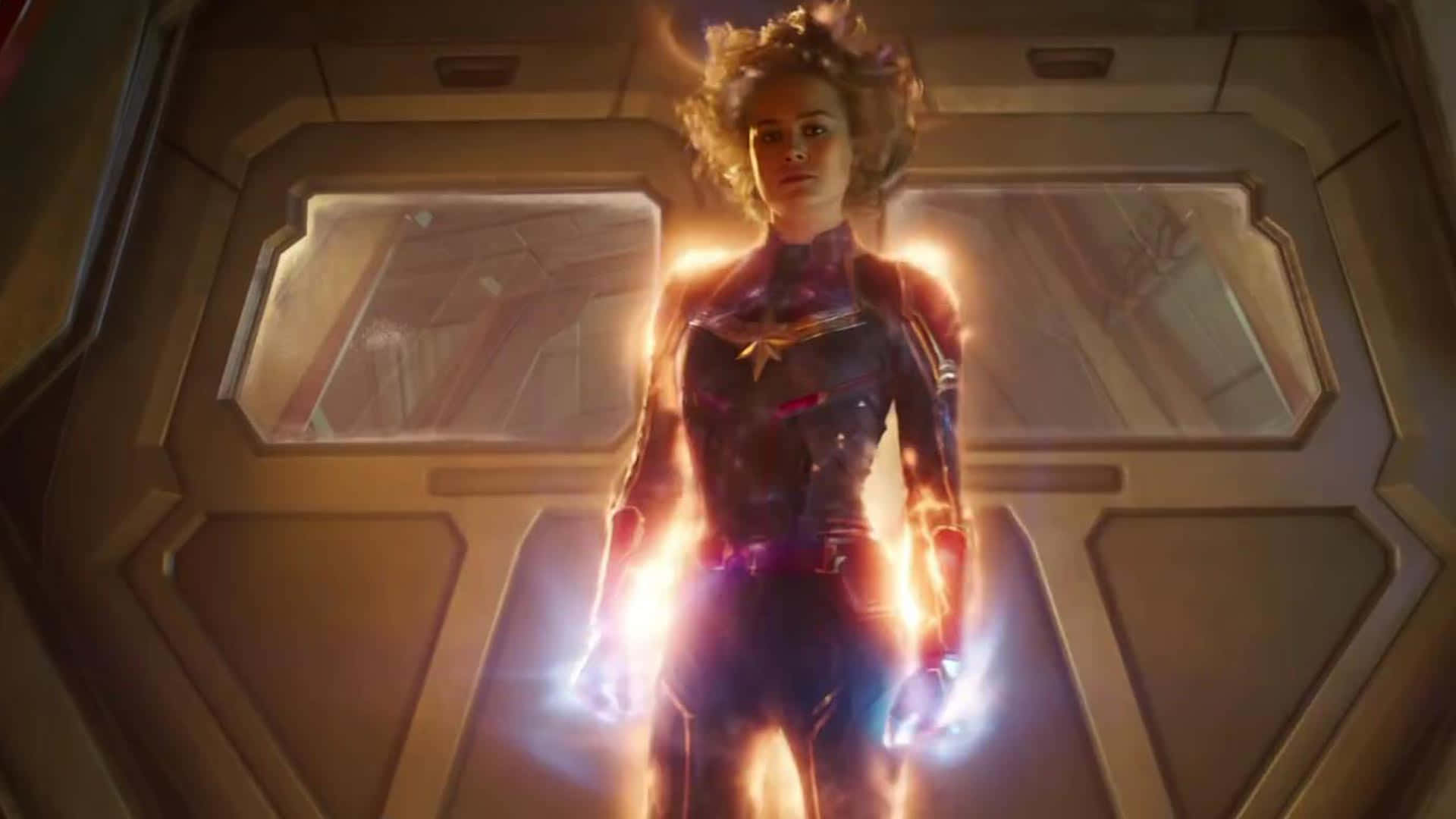 桌布 壁紙 — Captain Marvel 站在一條未來風格的太空船走廊中，雙手和頭髮散發出耀眼的光芒。