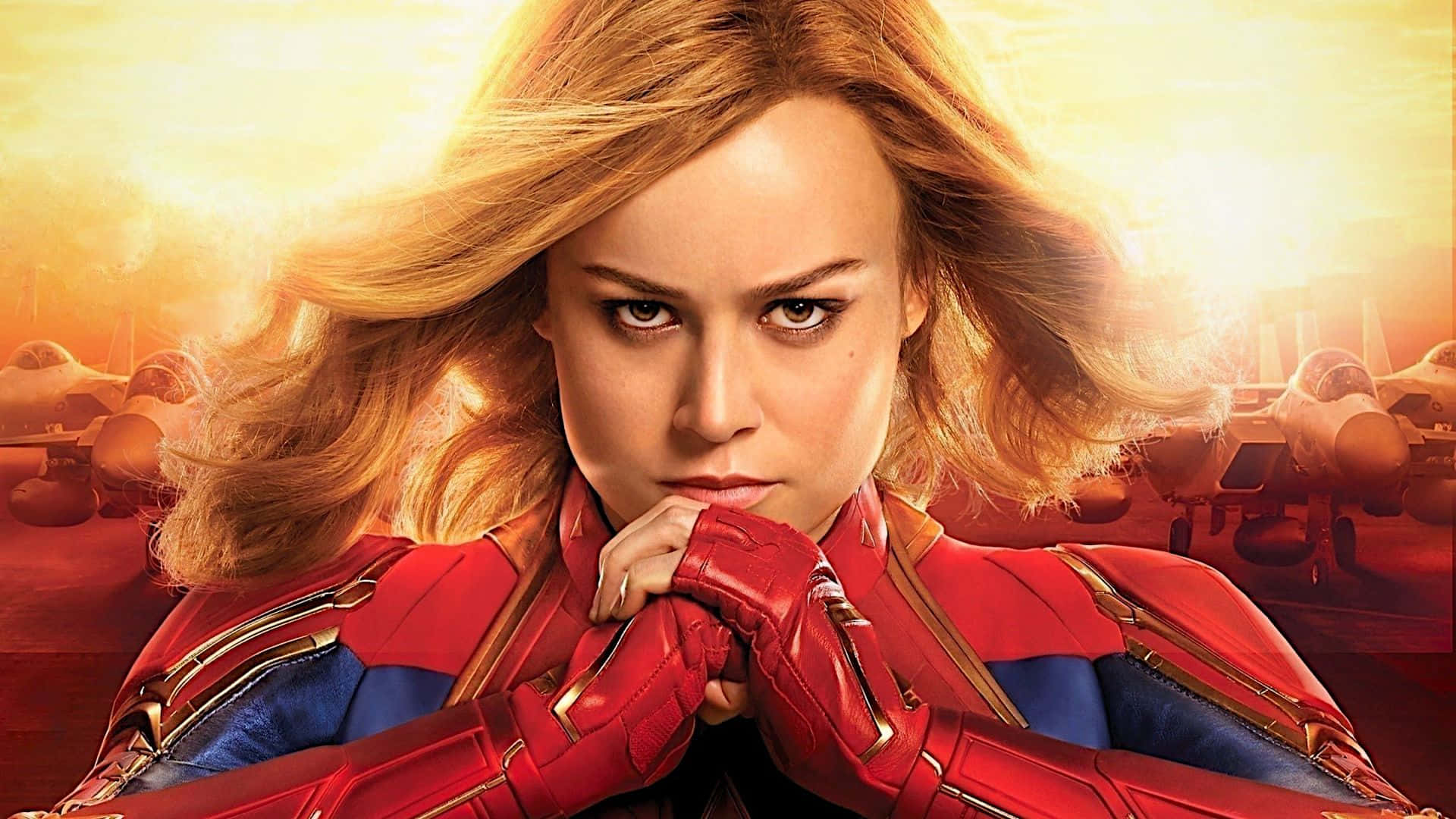 Captain Marvel HD 桌布 壁紙 — Captain Marvel 自信地穿著她的紅色和藍色套裝，身後有戰鬥機，背景是發光的日落天空。