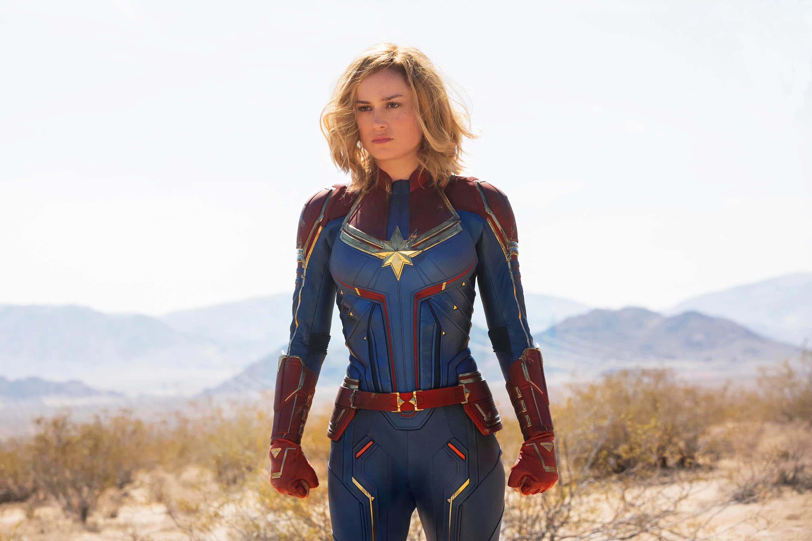 Captain Marvel 3d 桌布 壁紙 — Captain Marvel 自信地站在沙漠景觀中，穿著她標誌性的藍色和紅色服裝，帶有星星標誌。