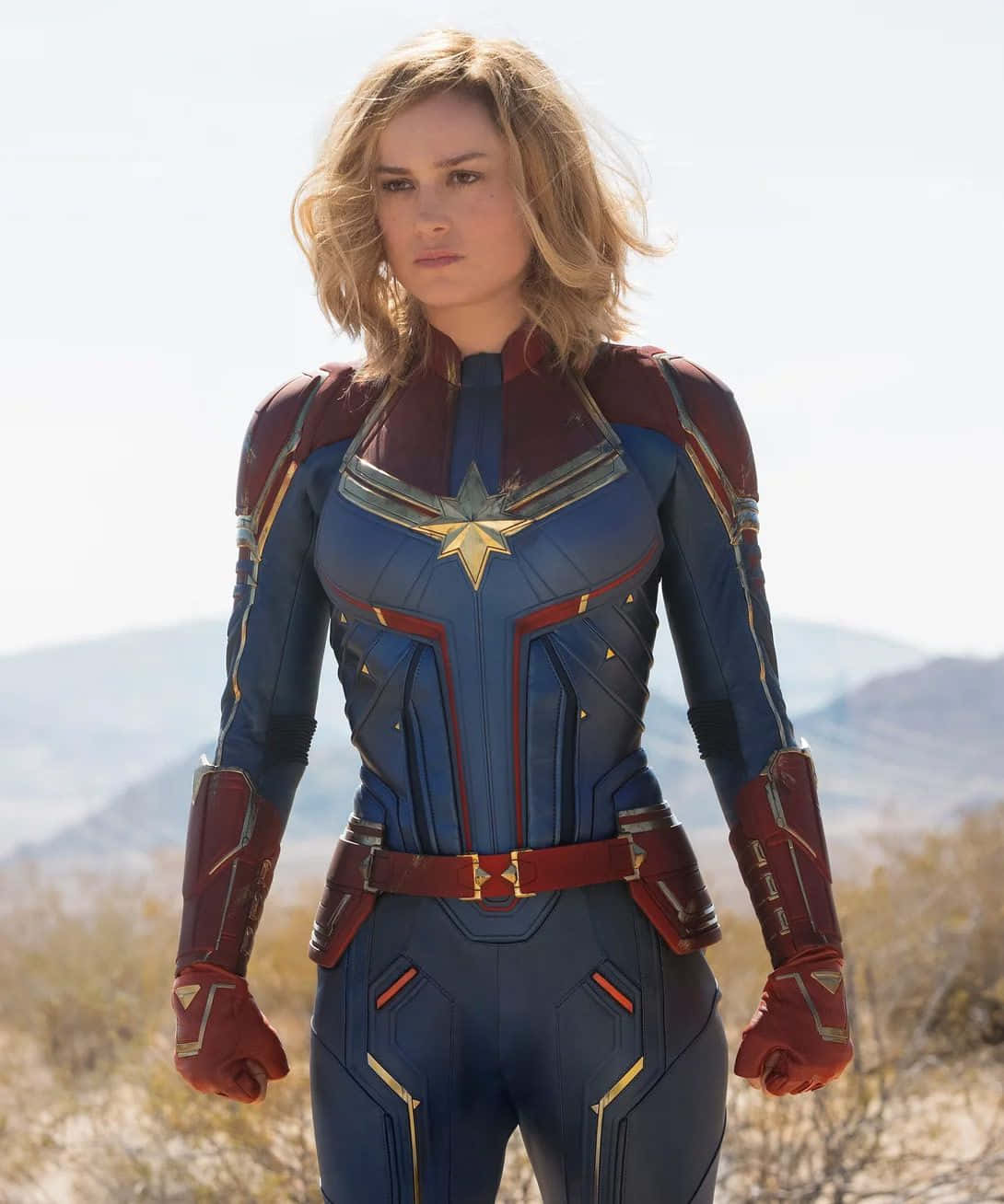 Captain Marvel 桌布 壁紙 — Captain Marvel 自信地穿著她的藍色和紅色戰衣，站在陽光明媚的沙漠背景下，遠方有山脈。