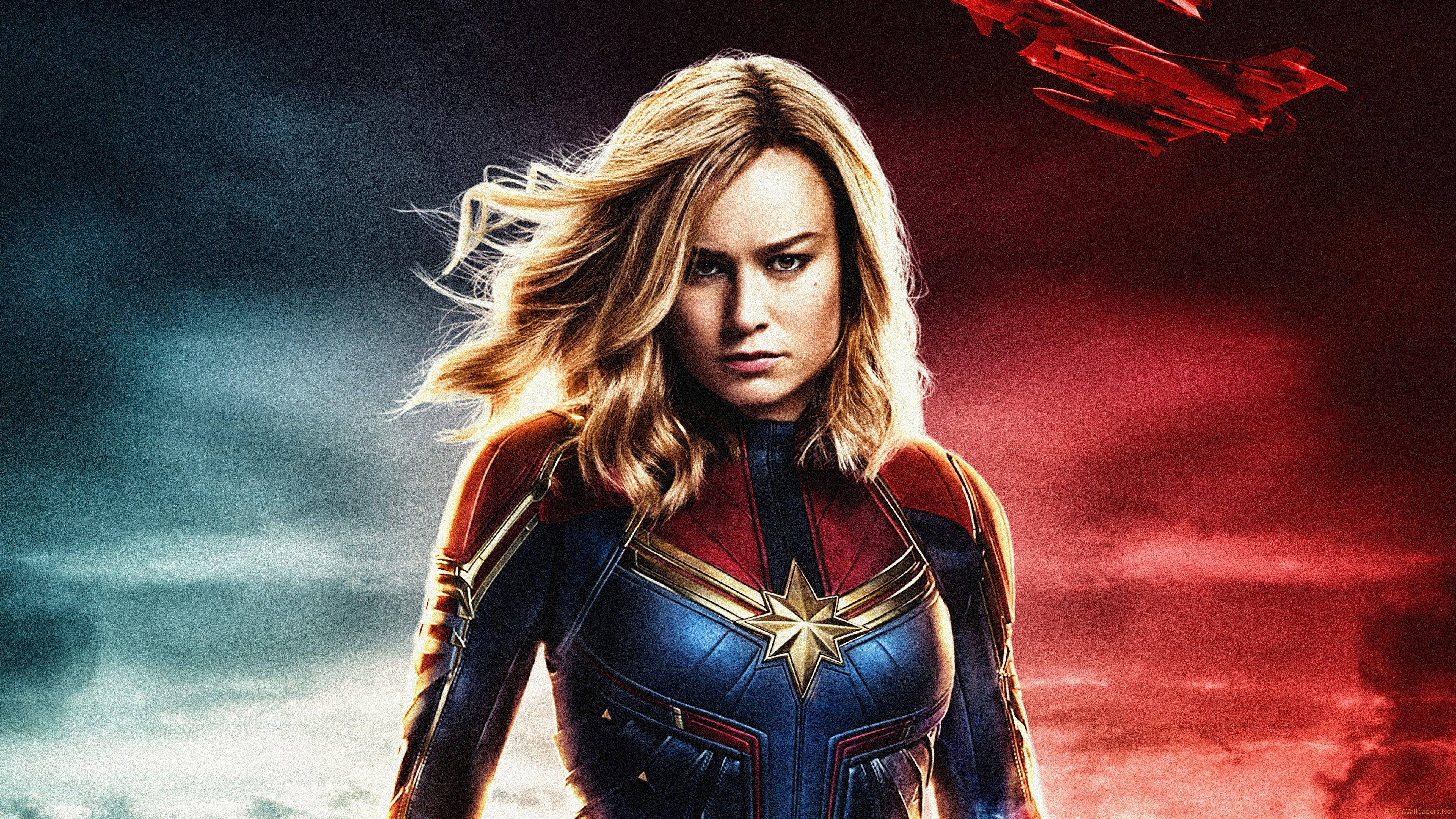 captain marvel 4K 桌布 壁紙 — Captain Marvel 身穿藍紅戰衣，在風暴雲和火紅色調劃分的戲劇性天空下堅定站立。