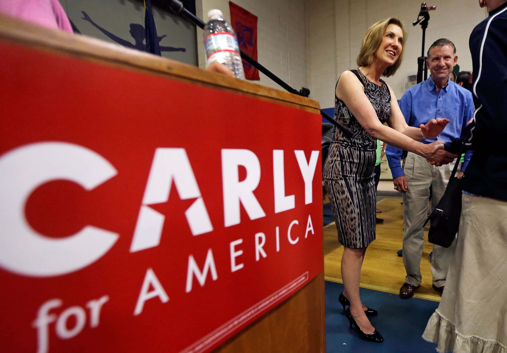 政治 桌布 壁紙 — Carly Fiorina 在競選活動中微笑握手，旁邊是紅色的「CARLY for AMERICA」講台。