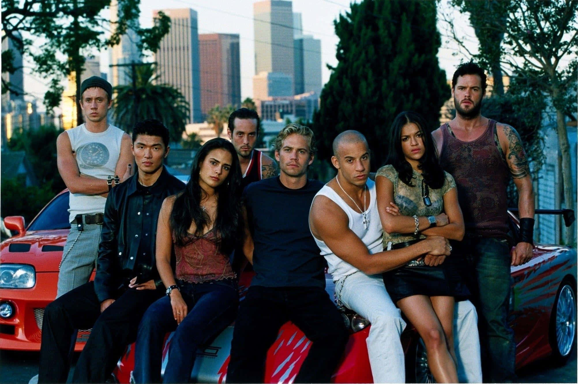 Fast And Furious 1 桌布 壁紙 — Cast of Fast and Furious 在日落時分的城市天際線背景下，自信地在一輛紅色跑車上擺姿勢。