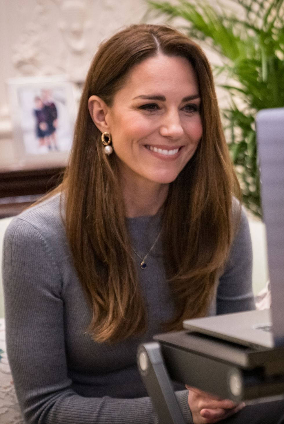 Kate Middleton 桌布 壁紙 — Catherine 在柔和燈光的室內環境中，坐在放有筆記型電腦的書桌前溫暖地微笑。