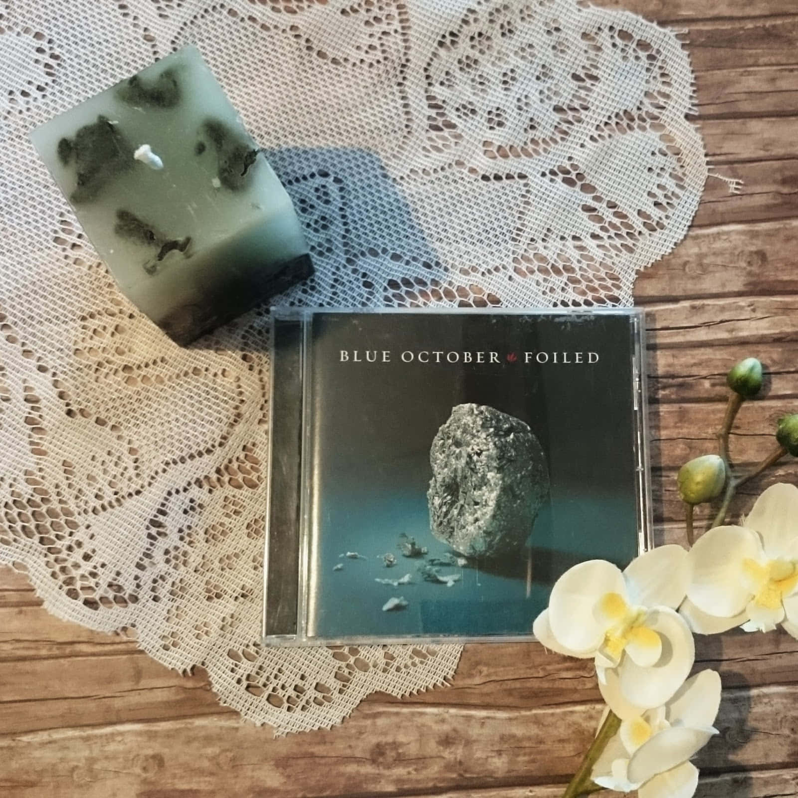 Blue October 桌布 壁紙 — Blue October's Foiled 的CD盒放在蕾絲上，旁邊有一塊綠色肥皂和白色蘭花在木質表面上。