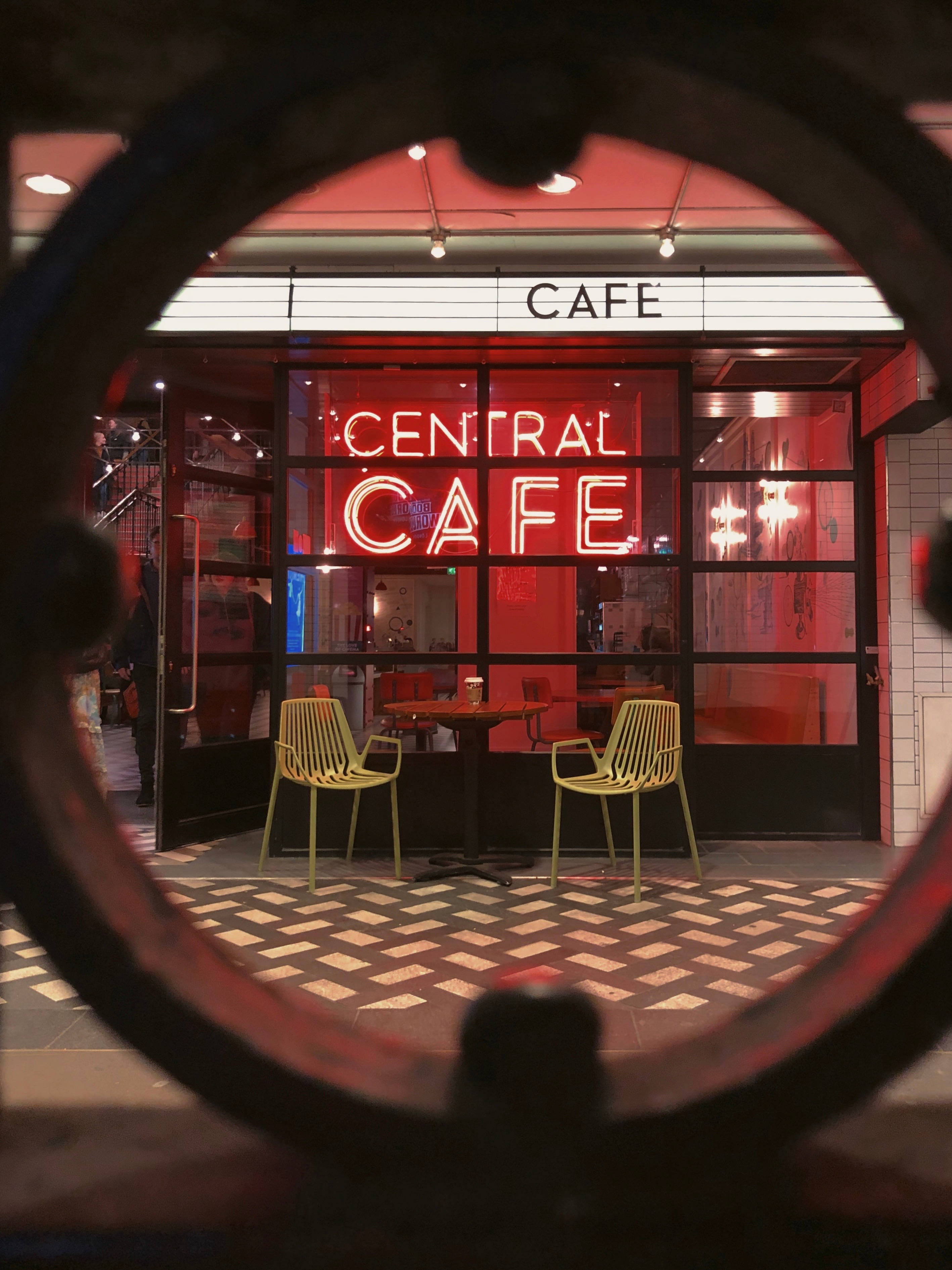 neon aesthetic iPhone 桌布 壁紙 — Central Cafe 的鮮紅色霓虹招牌透過圓形金屬框架發光，框架內是夜間棋盤格人行道上的兩張黃色椅子。