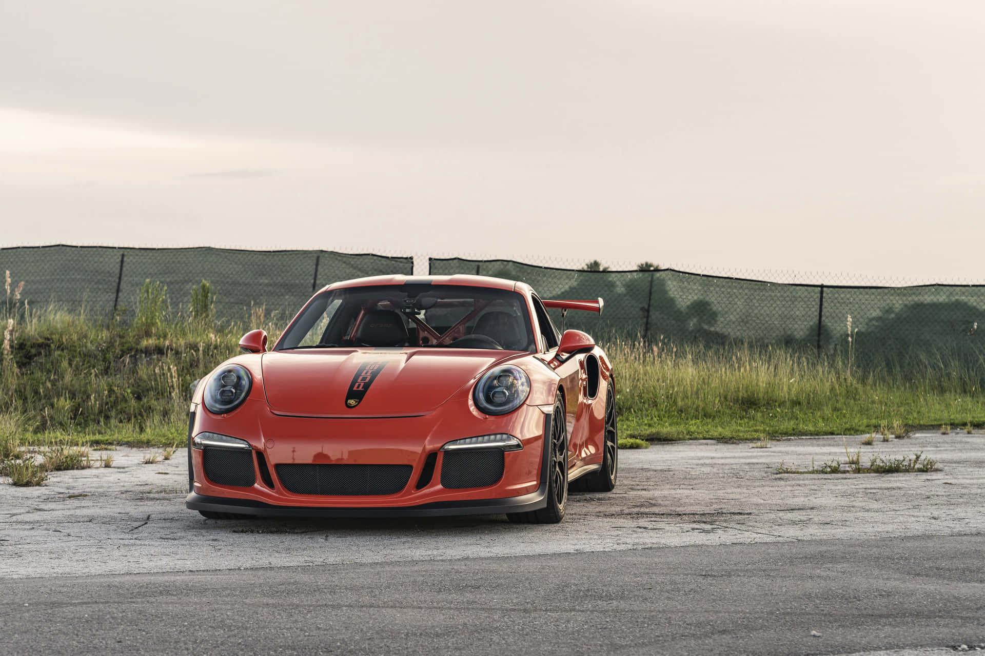  壁紙 — 一輛鮮豔的橙色保時捷911 GT3 RS跑車停在裂開的瀝青路上，後面有鐵絲網圍欄和雜草叢生的草地，天空朦朧。