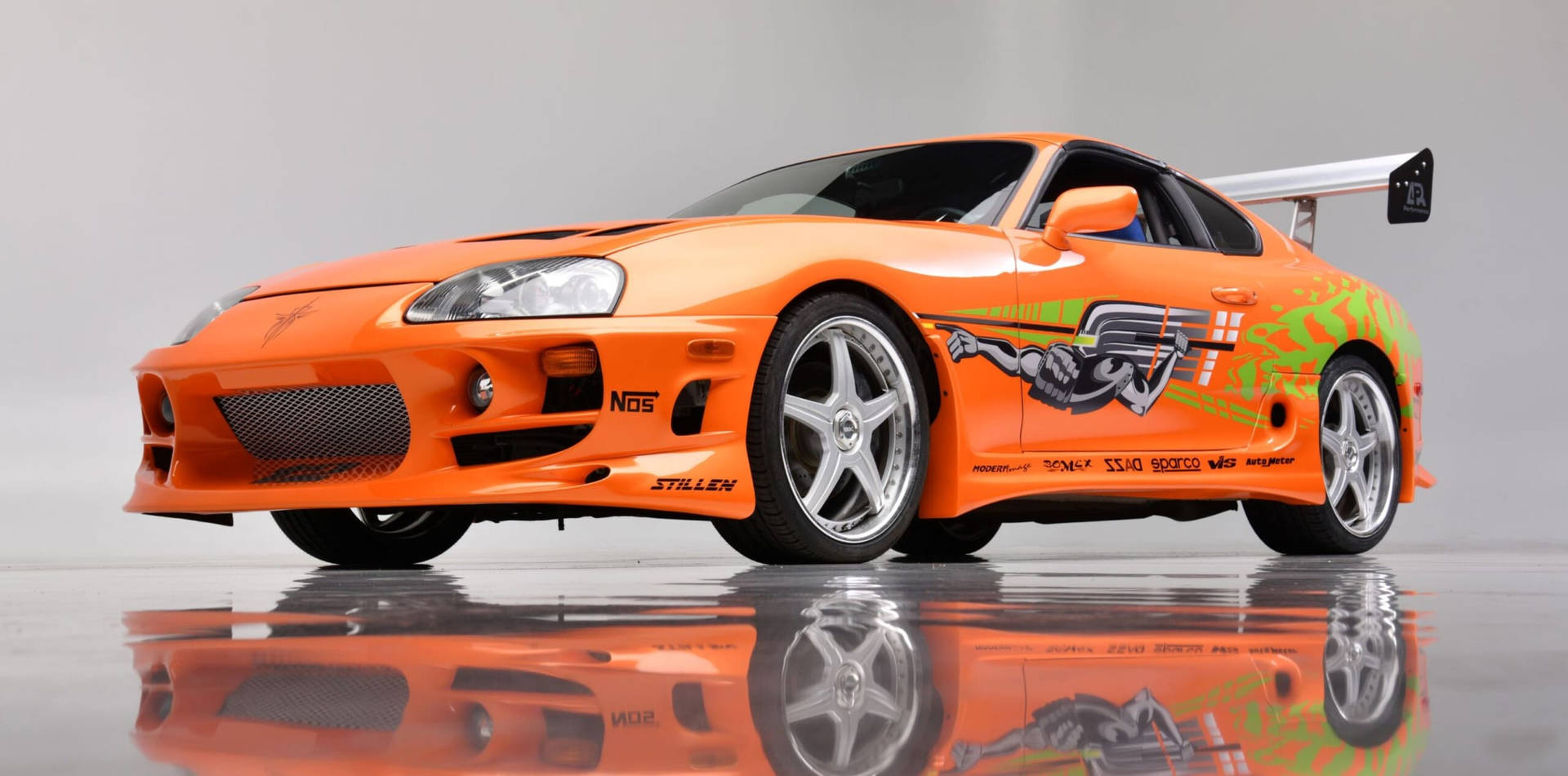 橙色豐田Supra，帶有賽車貼紙和後翼，在反射地板上。 — 保羅·沃克汽車 壁紙