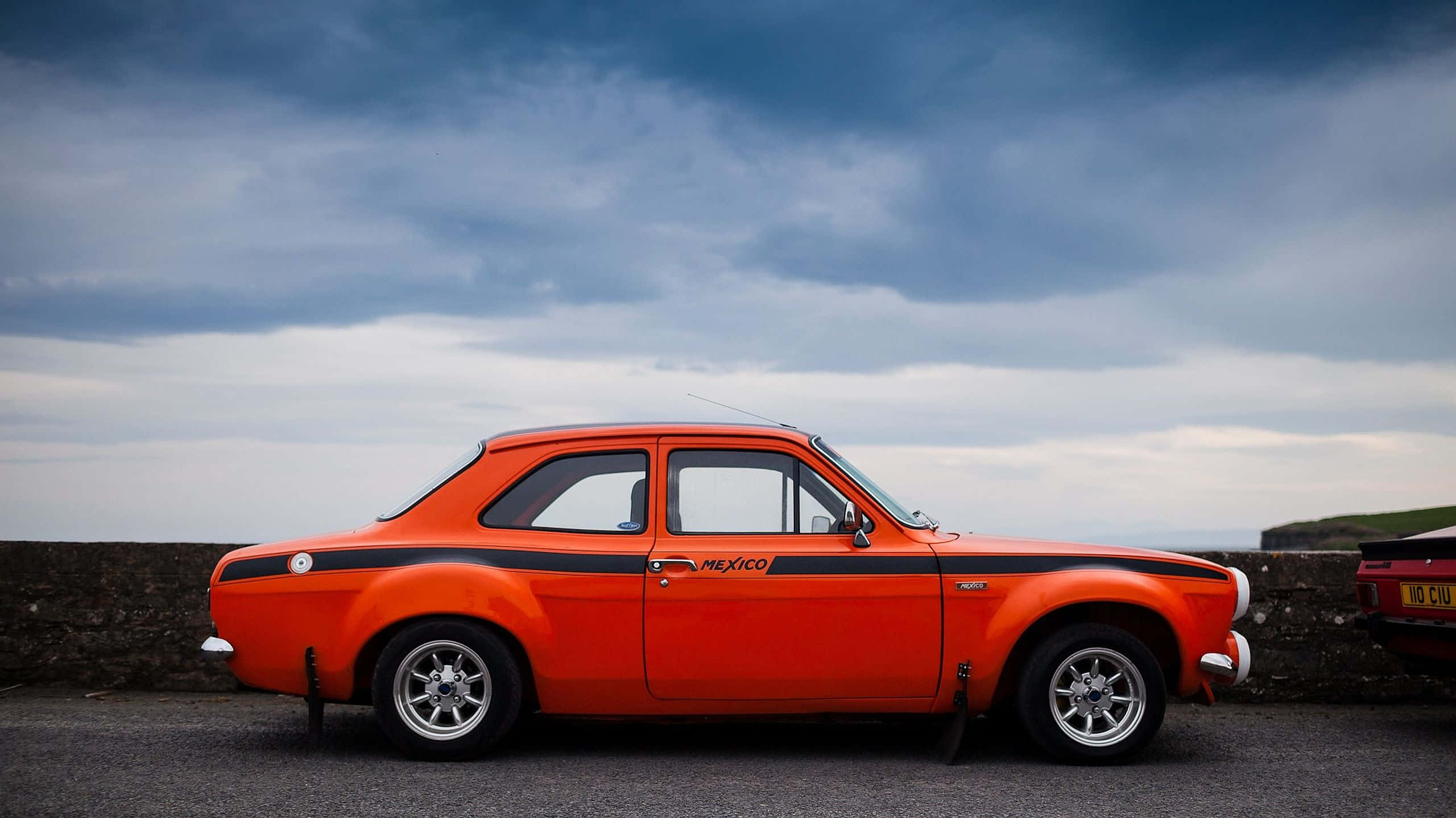 Ford Escort 桌布 壁紙 — 一輛充滿活力的橙色復古跑車，帶有黑色賽車條紋，停泊在海岸公路上，天空多雲而戲劇化。
