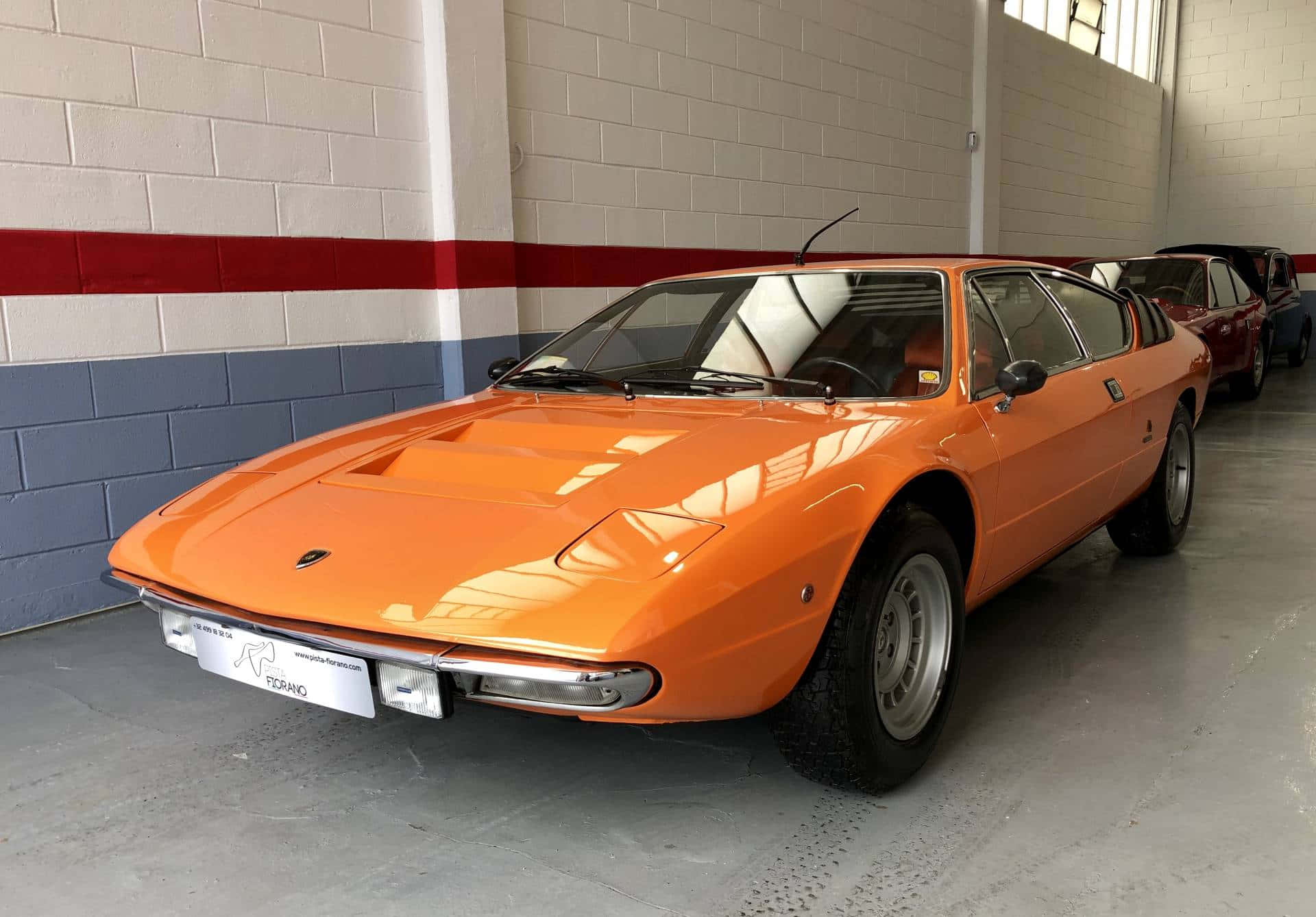 Lamborghini Urraco 桌布 壁紙 — 一輛充滿活力的橙色復古跑車在乾淨的室內車庫中，在明亮的燈光下閃閃發光，車庫有紅色和藍色的磚牆。