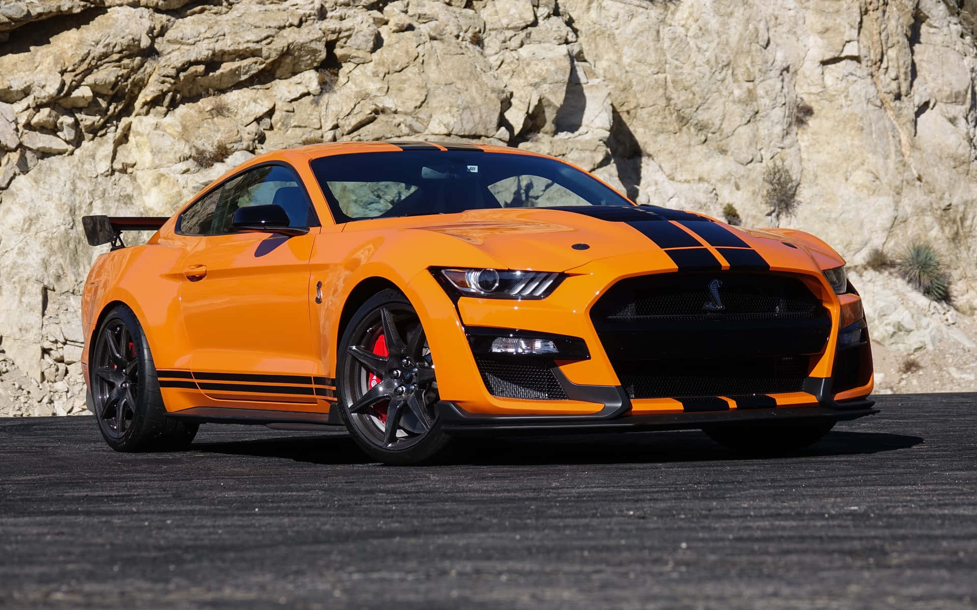  壁紙 — 一輛充滿活力的橙色福特野馬Shelby GT500，帶有黑色賽車條紋，停在瀝青路面上，背景是崎嶇的岩石懸崖，在明亮的日光下。