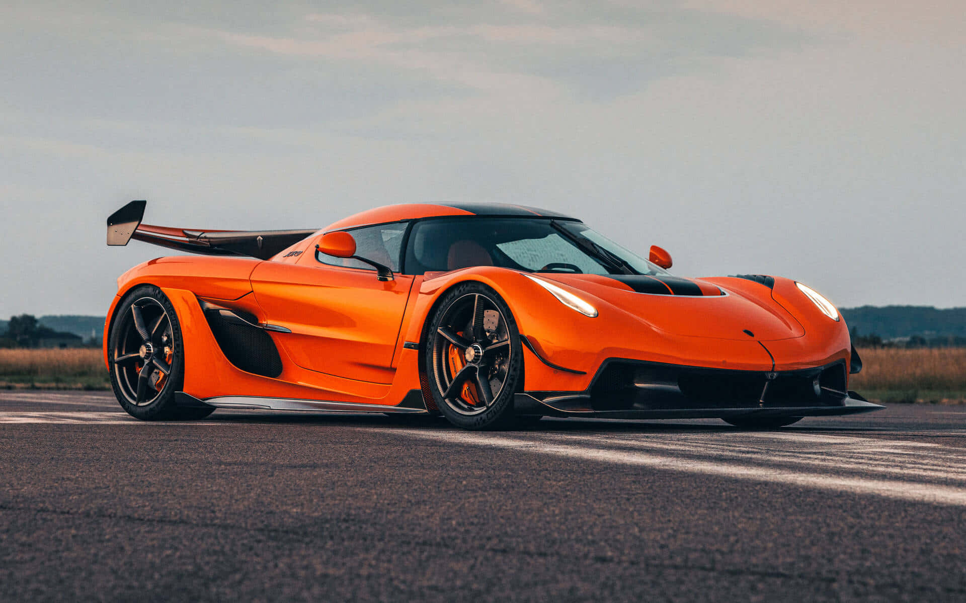  壁紙 — 一輛充滿活力的橙色 Koenigsegg 超級跑車，帶有黑色飾邊和大型後擾流板，停在黃昏的瀝青跑道上。