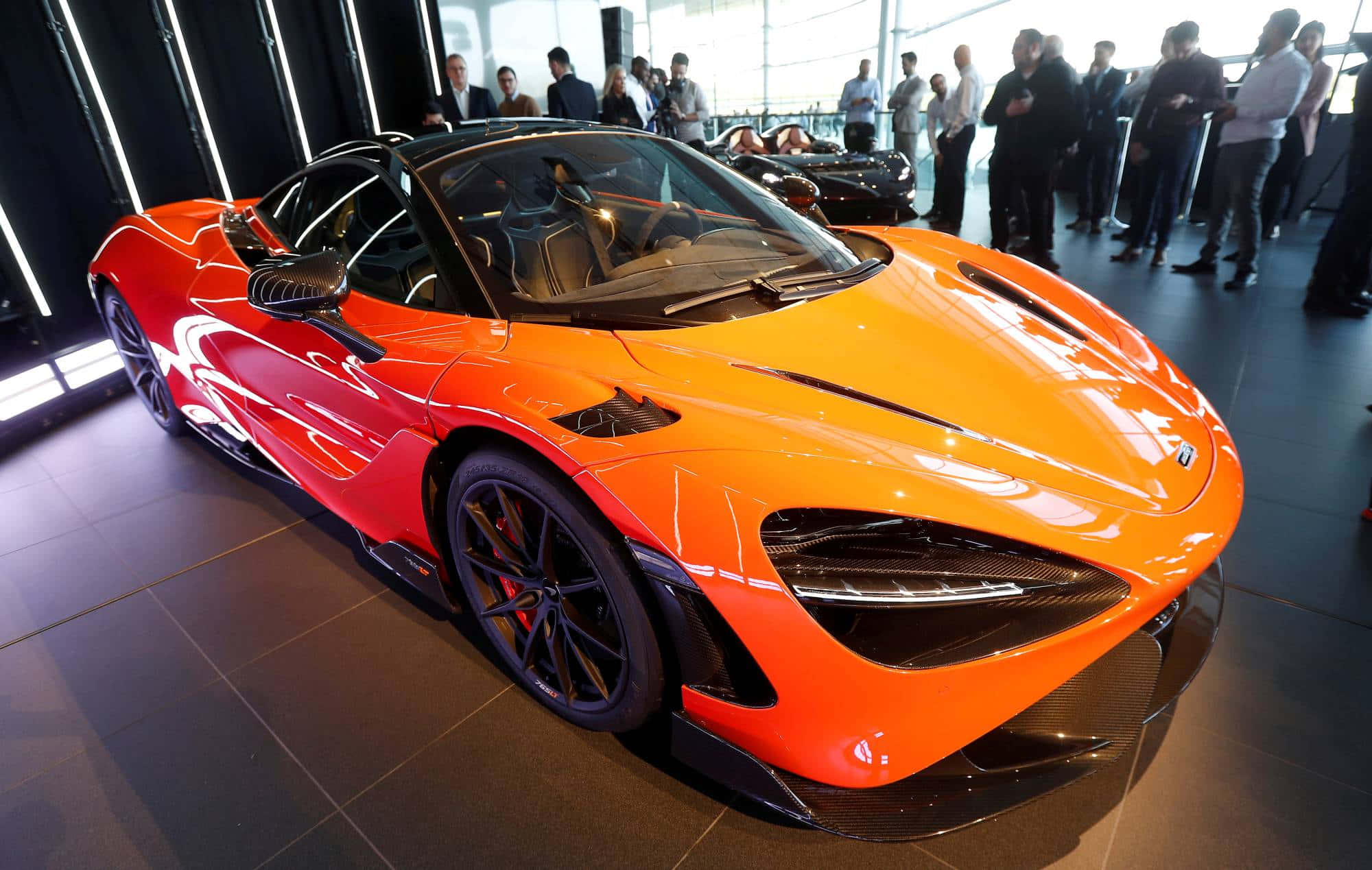 Mclaren 765lt 桌布 壁紙 — 一輛充滿活力的橙色McLaren超級跑車在明亮燈光下閃閃發光，觀眾聚集在時尚現代展廳周圍。