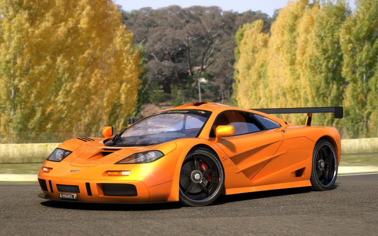 Mclaren F1 桌布 壁紙 — 一輛鮮豔的橙色McLaren F1跑車在金色秋樹環繞的道路上，在晴朗的日光下閃閃發光。