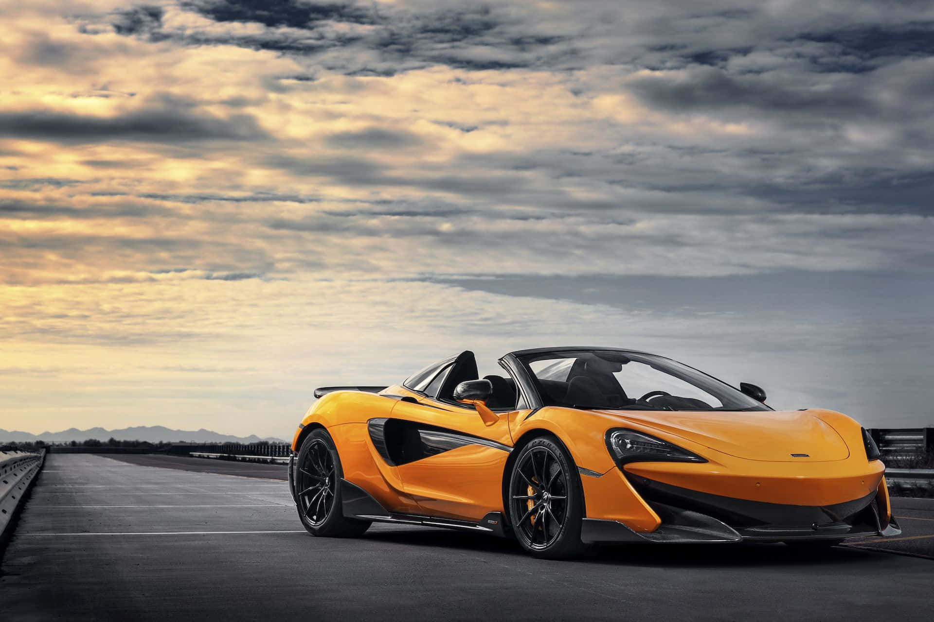 Mclaren 600lt 桌布 壁紙 — 一輛充滿活力的橙色McLaren跑車，帶有黑色飾邊，停在一條長路上，背景是日落時分戲劇性的多雲天空。