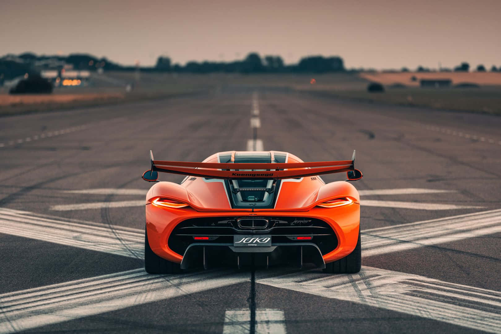  壁紙 — 一輛鮮豔的橙色McLaren Senna跑車在黃昏時分停放在寬闊的瀝青跑道上，尾燈閃爍，天空壯麗。