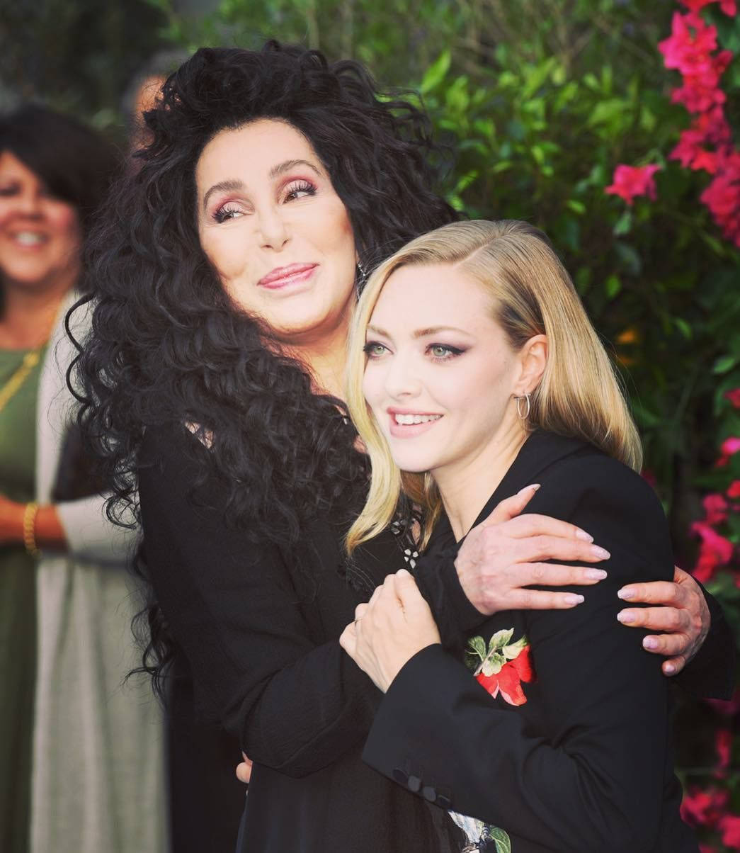 Amanda Seyfried 桌布 壁紙 — Cher 在戶外溫暖地擁抱Amanda Seyfried，周圍是茂密的綠色植物和鮮豔的粉紅色花朵。