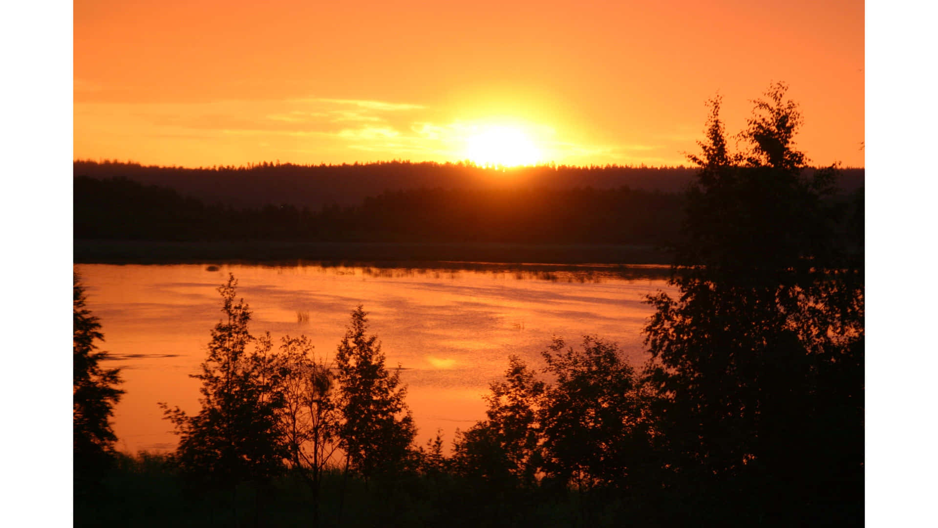 壁紙 — 熾熱的夕陽在平靜的湖面上投射出溫暖的橙色光芒，襯托出樹木的剪影和遠處的森林山丘。