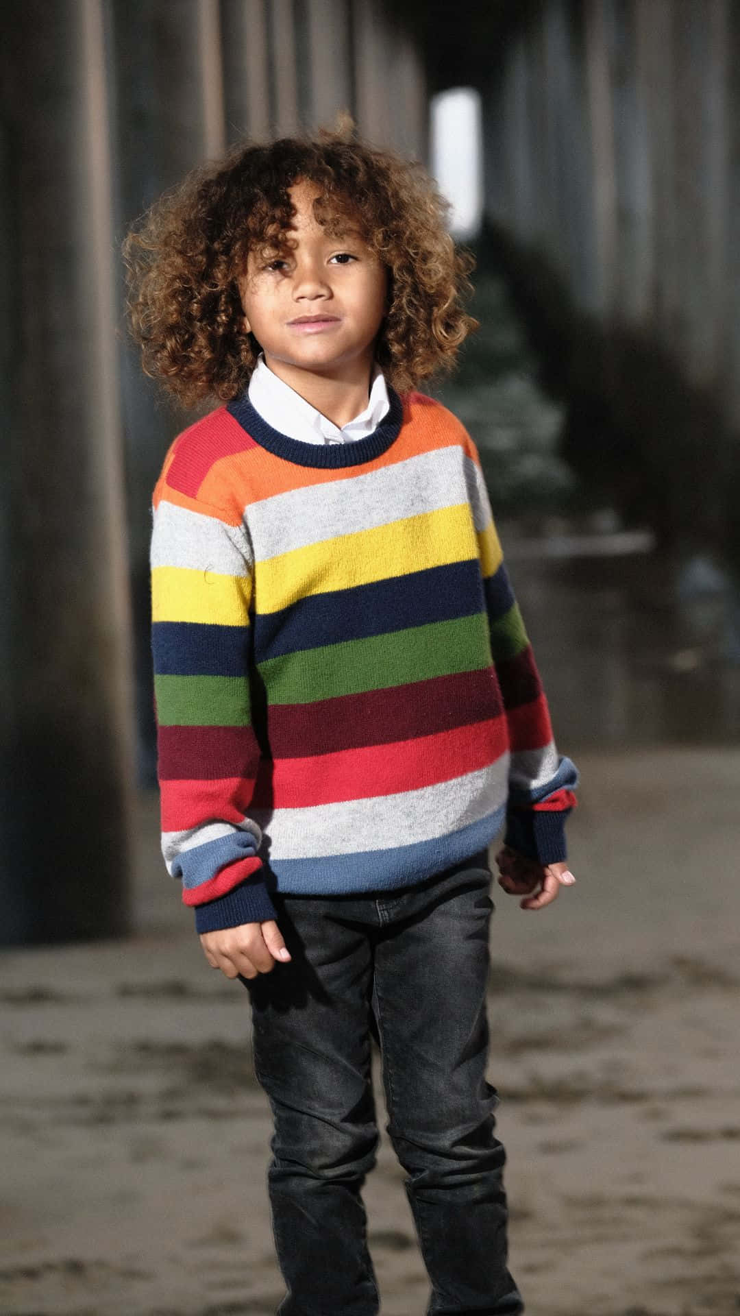  壁紙 — 一個捲髮小孩自信地穿著彩色條紋毛衣站在暮色中的碼頭下。