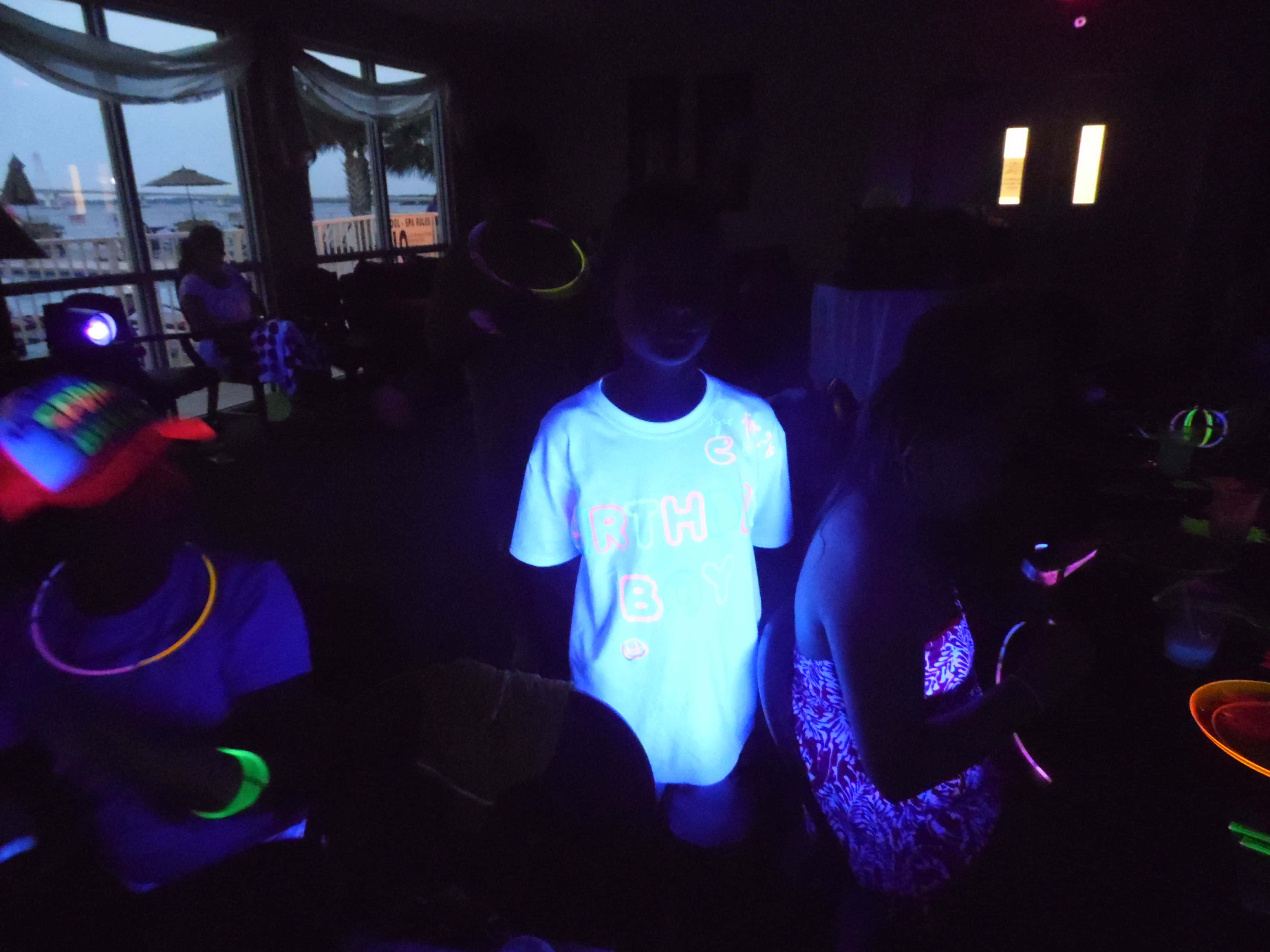 Neon Party 桌布 壁紙 — 兒童在黑暗房間中佩戴霓虹配件，在黑光下發光，窗外可見海灘。