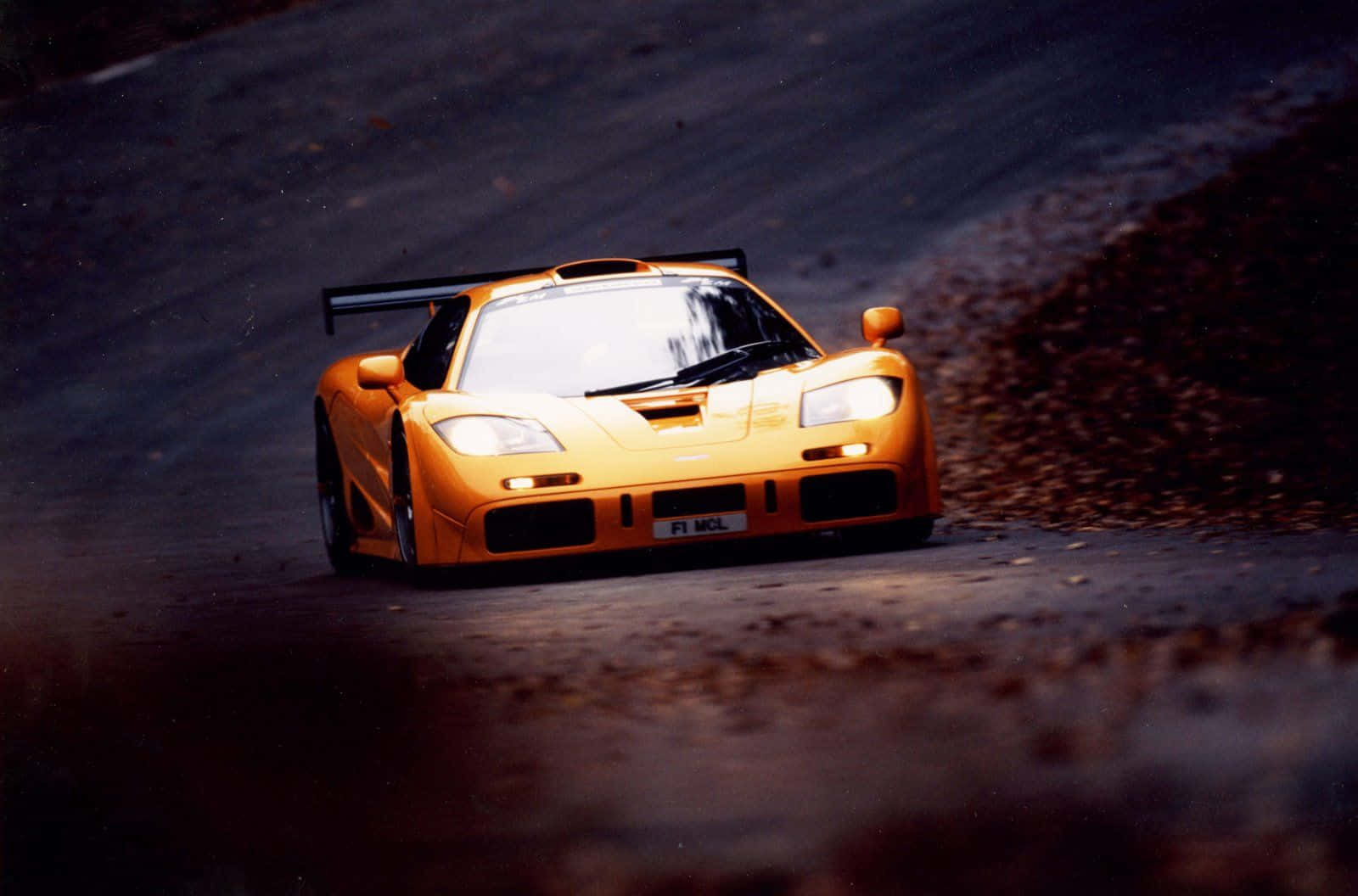Mclaren F1 桌布 壁紙 — 一輛充滿活力的橙色McLaren F1賽車在黃昏時分沿著黑暗蜿蜒的道路疾馳，帶有動態模糊和戲劇性燈光。