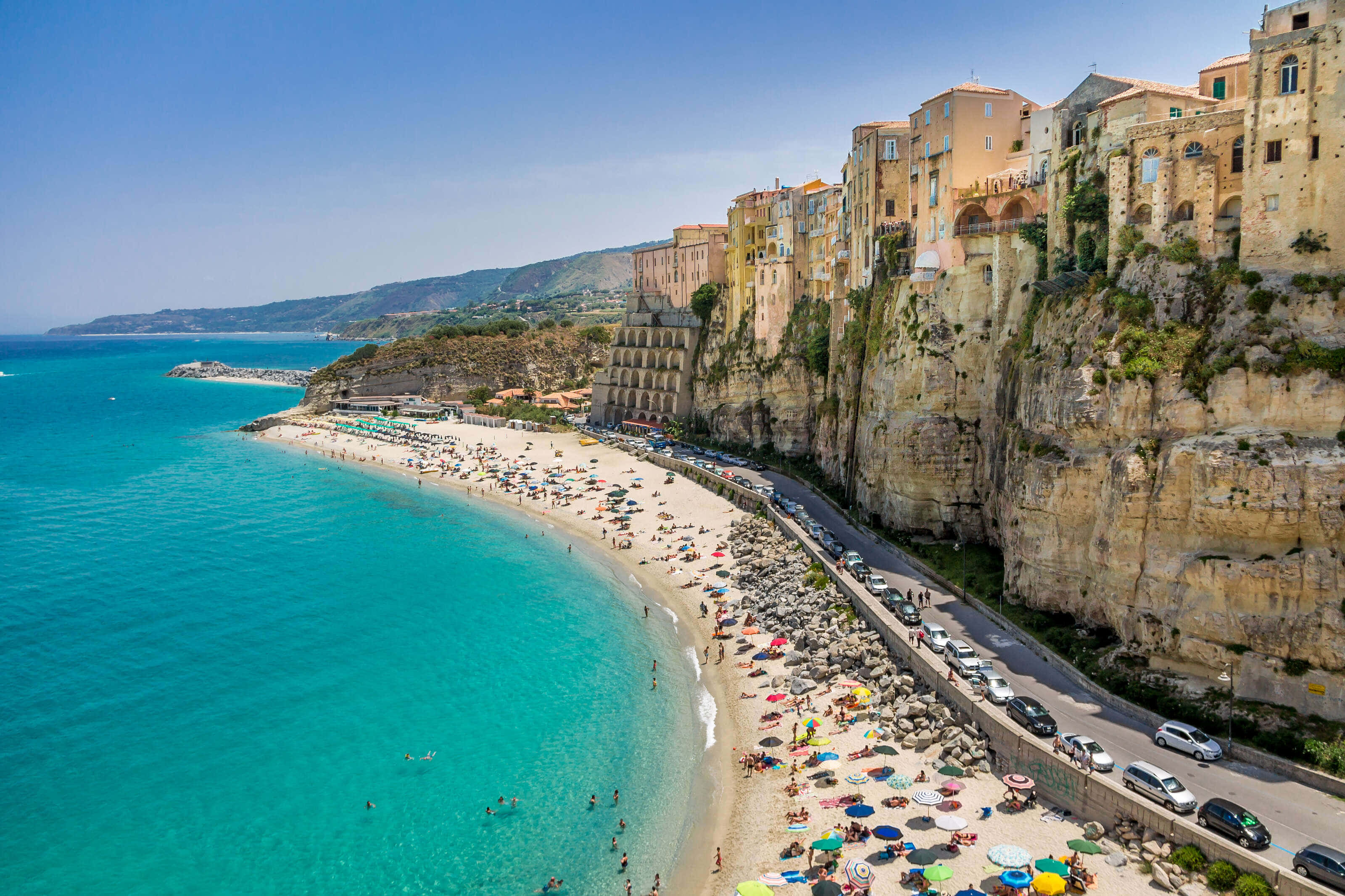 Italian Beach 桌布 壁紙 — 充滿活力的藍綠色海水拍打著繁忙的沙灘，沙灘下方是歷史悠久的懸崖建築，上方是清澈的藍天。