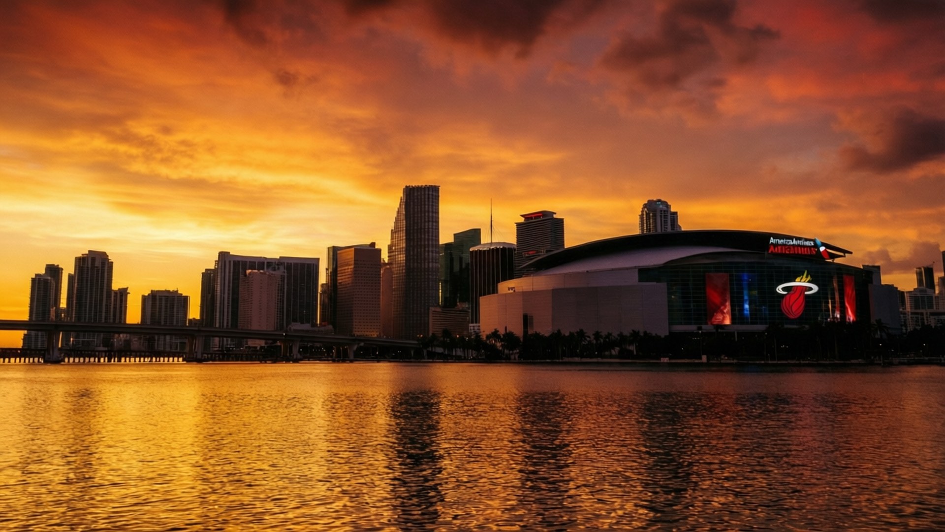 Miami Sunset Arena 壁紙 — 充滿活力的日落將火熱的橙色和金色色調投射在邁阿密的天際線上，美國航空競技場在平靜的水域旁格外突出。