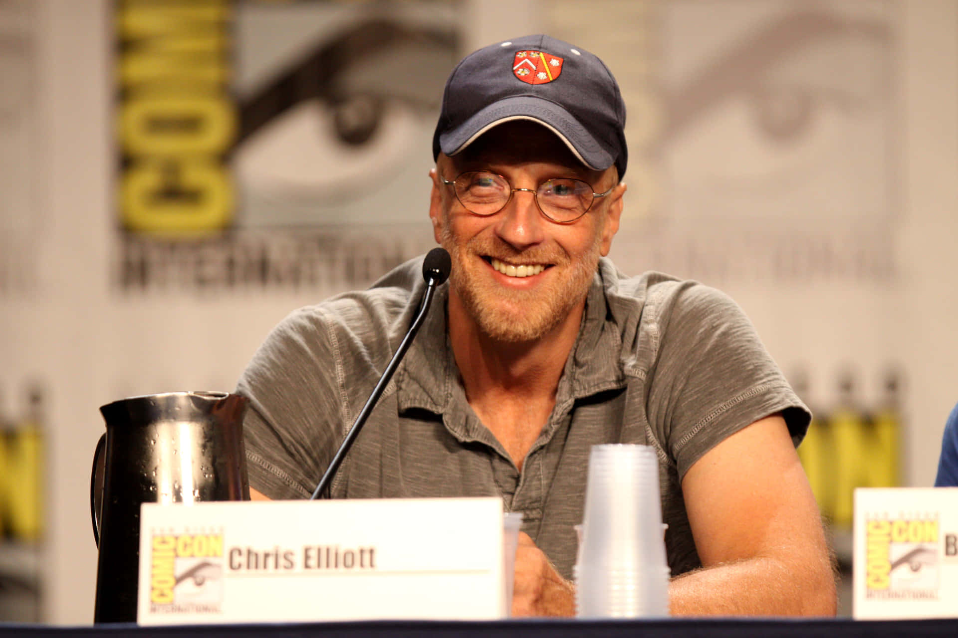  壁紙 — Chris Elliott 在Comic-Con座談會上戴著帽子和眼鏡，面前有麥克風，溫暖地微笑。