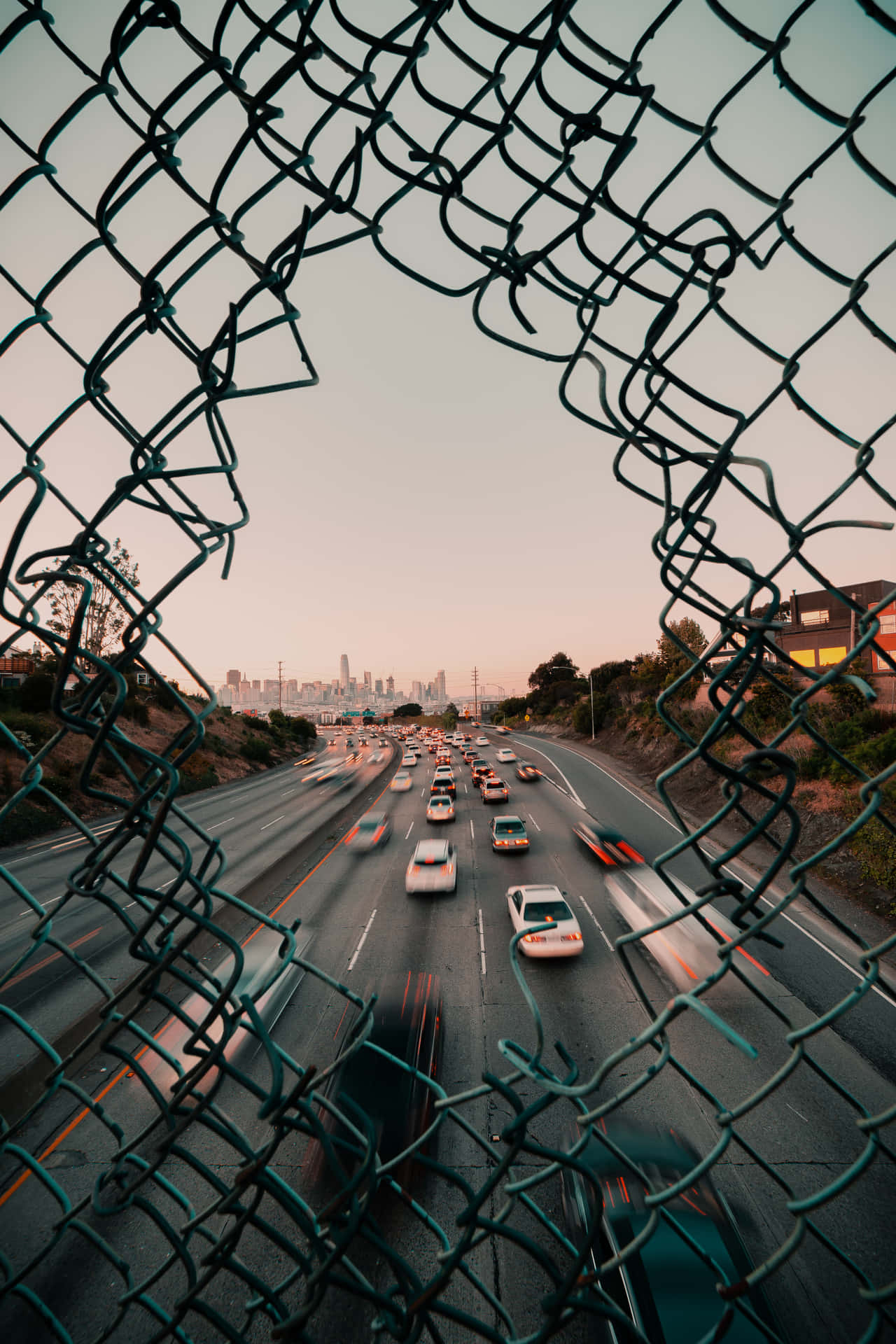 穿過鐵絲網圍欄看到的繁忙高速公路，汽車在黃昏時分行駛。 — iPad 壁紙