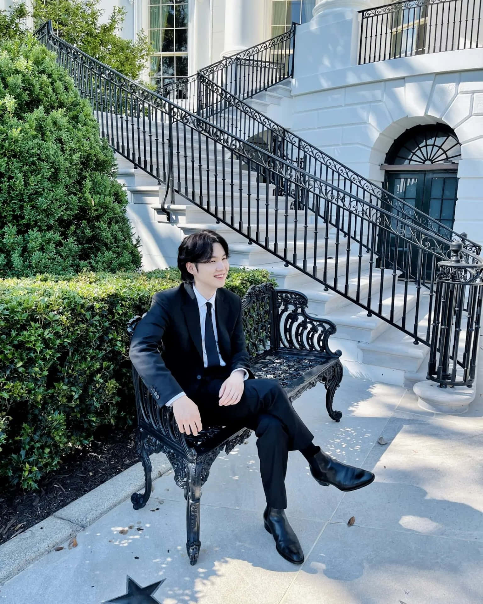  壁紙 — 一名身穿俐落黑色西裝的男子，放鬆地坐在一座宏偉白色大宅前裝飾華麗的長椅上，大宅有寬闊的樓梯和茂密的綠色樹籬。