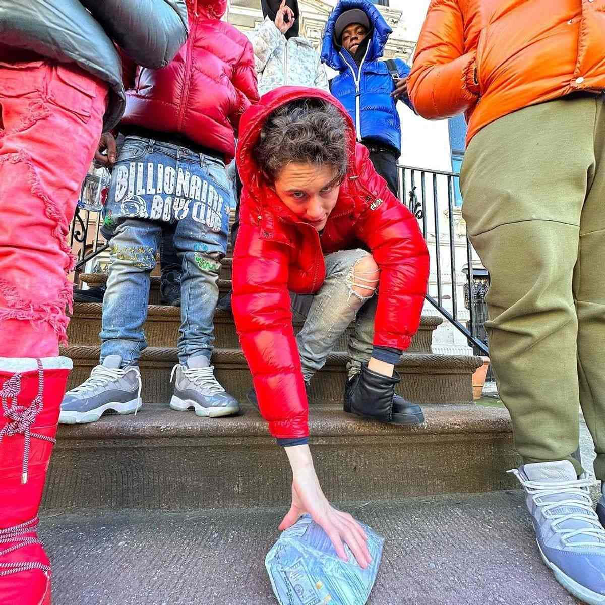  壁紙 — 一個穿著紅色羽絨外套的年輕人蹲在石階上，伸手去拿一疊現金，周圍是其他穿著色彩繽紛街頭服飾的人。