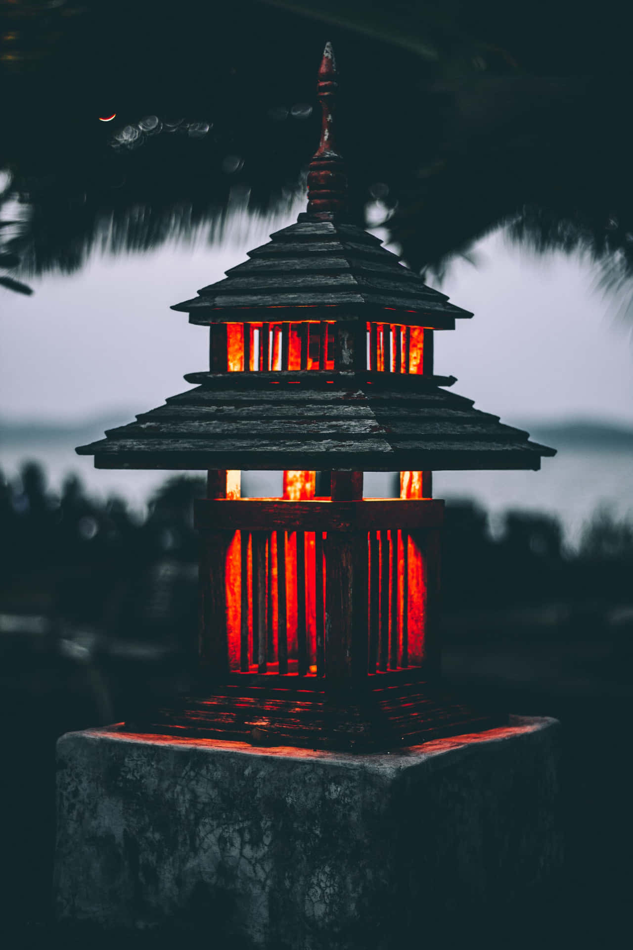 一個發光的紅色木燈籠，帶有分層屋頂，在黃昏時立於石基上。 — 日式暗黑 壁紙