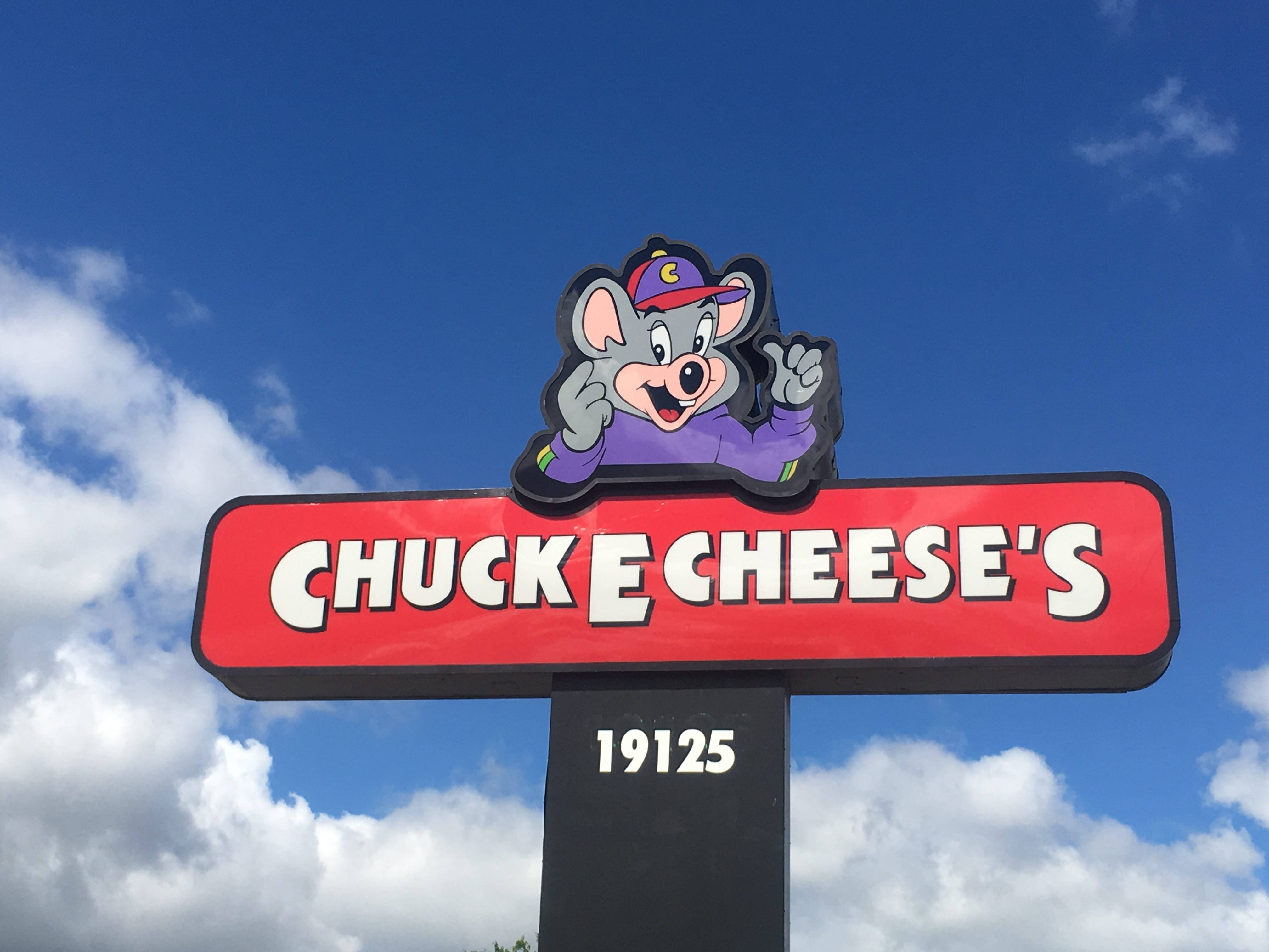 E 桌布 壁紙 — Chuck E Cheese 的吉祥物標誌，卡通老鼠在明亮的藍天和蓬鬆的白雲下揮手。