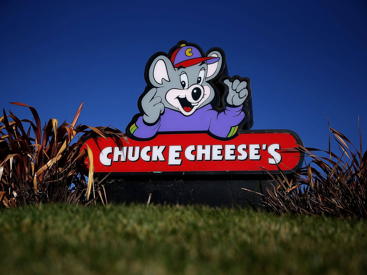 E 桌布 壁紙 — Chuck E Cheese 的吉祥物標誌，卡通老鼠揮手站在草地上，背景是晴朗藍天。