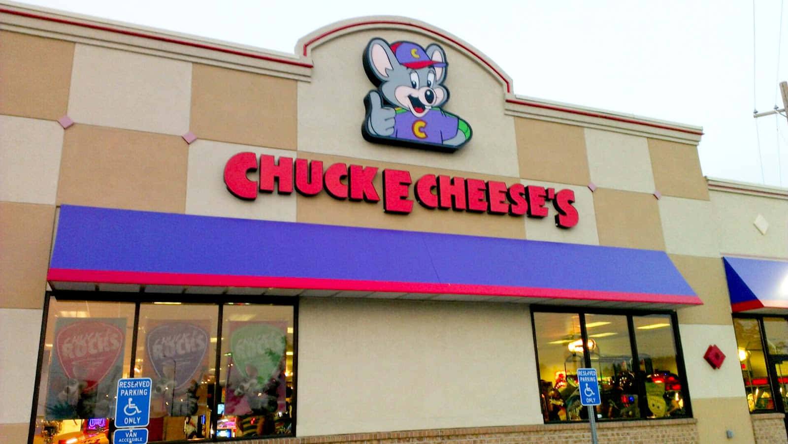 查克芝士 桌布 壁紙 — Chuck E Cheese's 的店面，有卡通老鼠吉祥物在紫色遮陽篷下的紅色標誌上方豎起大拇指。