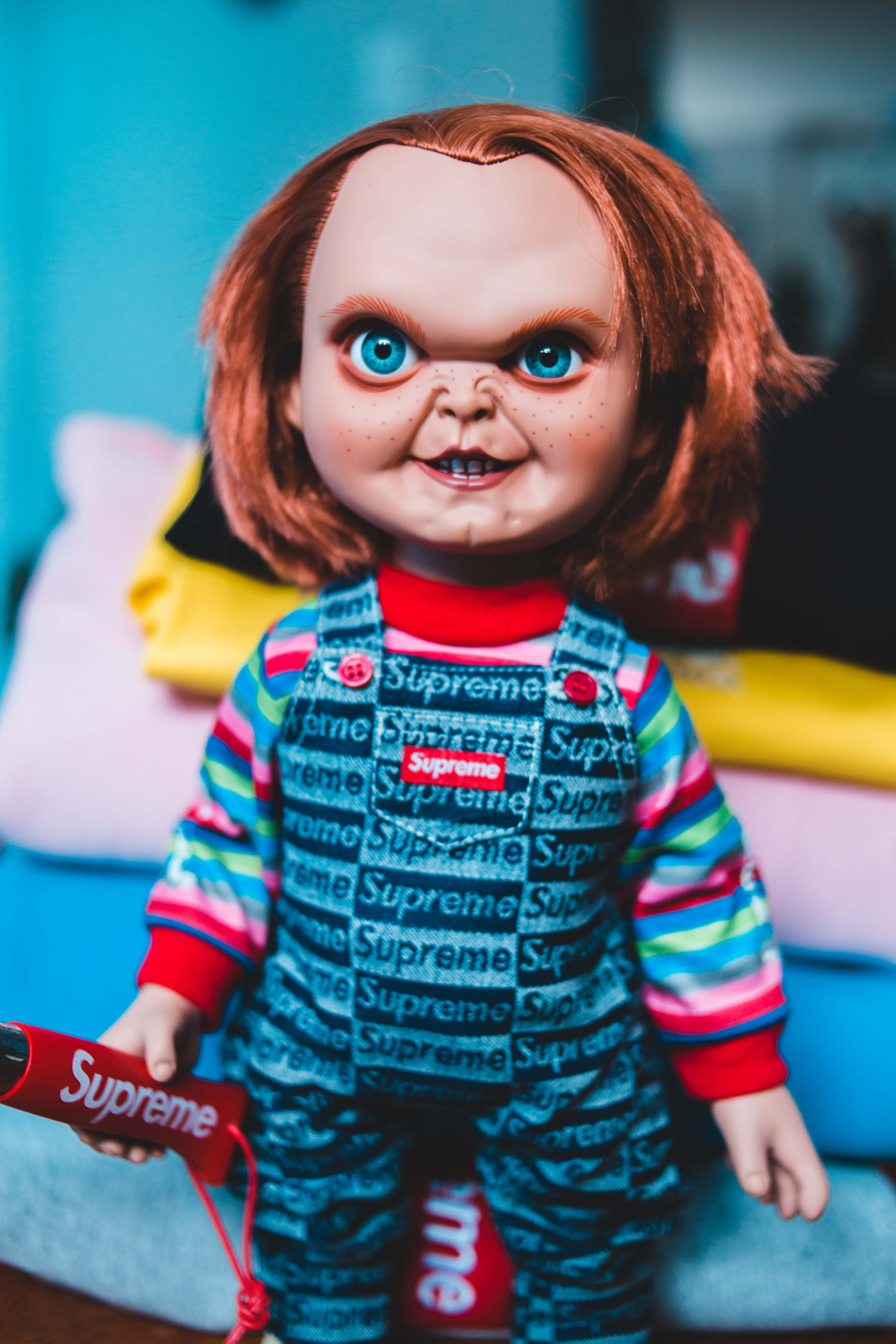 鬼娃恰吉 桌布 壁紙 — Chucky 公仔的近距離特寫，穿著 Supreme 品牌服裝，擁有明亮的藍色眼睛和紅色頭髮，背景是模糊的彩色。