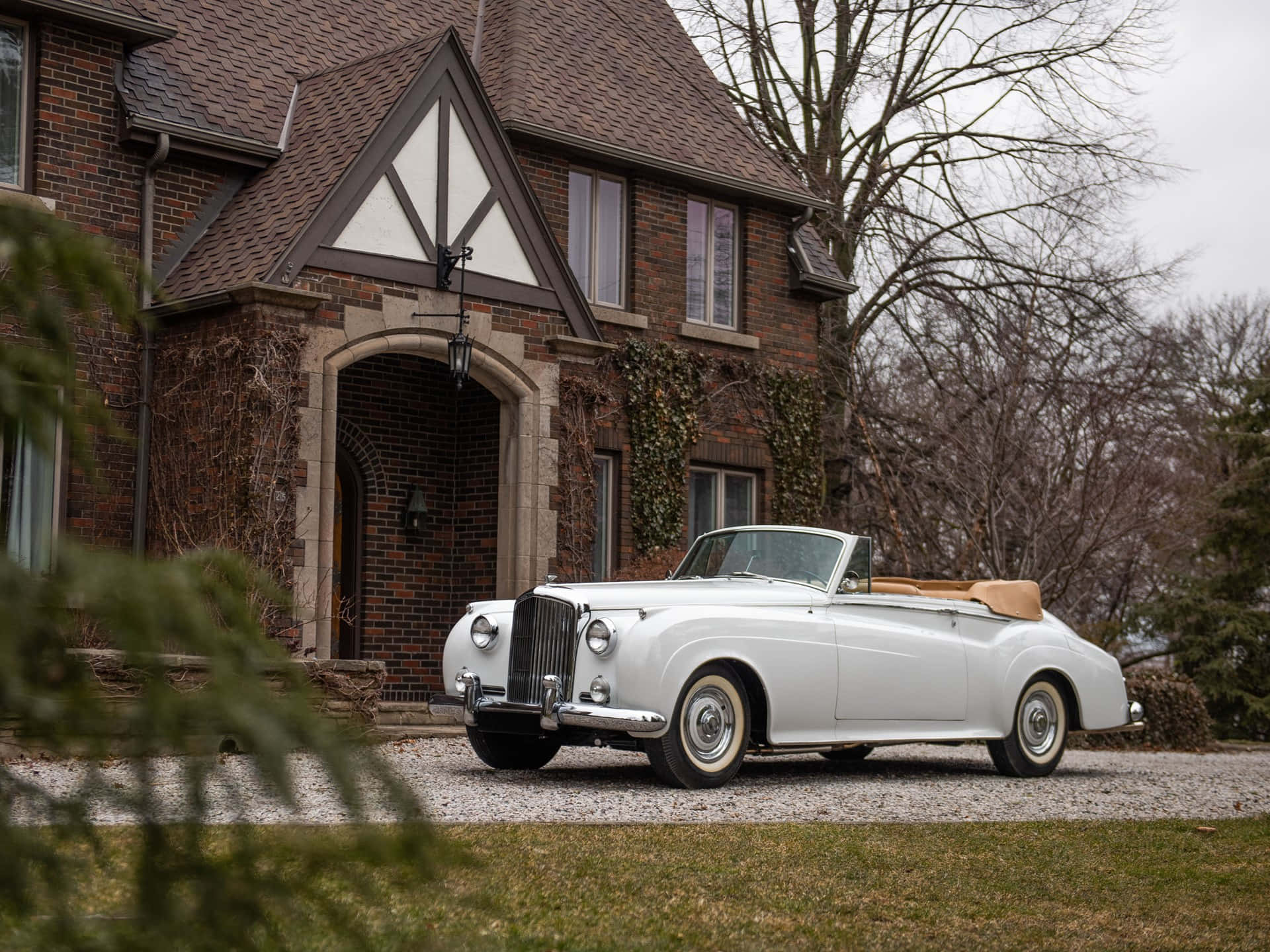 Bentley S2 桌布 壁紙 — 一輛純白色復古敞篷車停在碎石路上，前面是一座莊嚴的磚砌都鐸式房屋，周圍是光禿禿的冬季樹木。