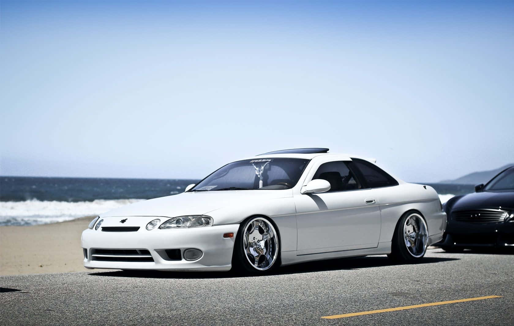 Lexus Sc 300 桌布 壁紙 — 一輛純白色運動型轎跑車，配備定制鍍鉻輪圈，停泊在沿海公路上，旁邊是海洋，天空清澈湛藍。
