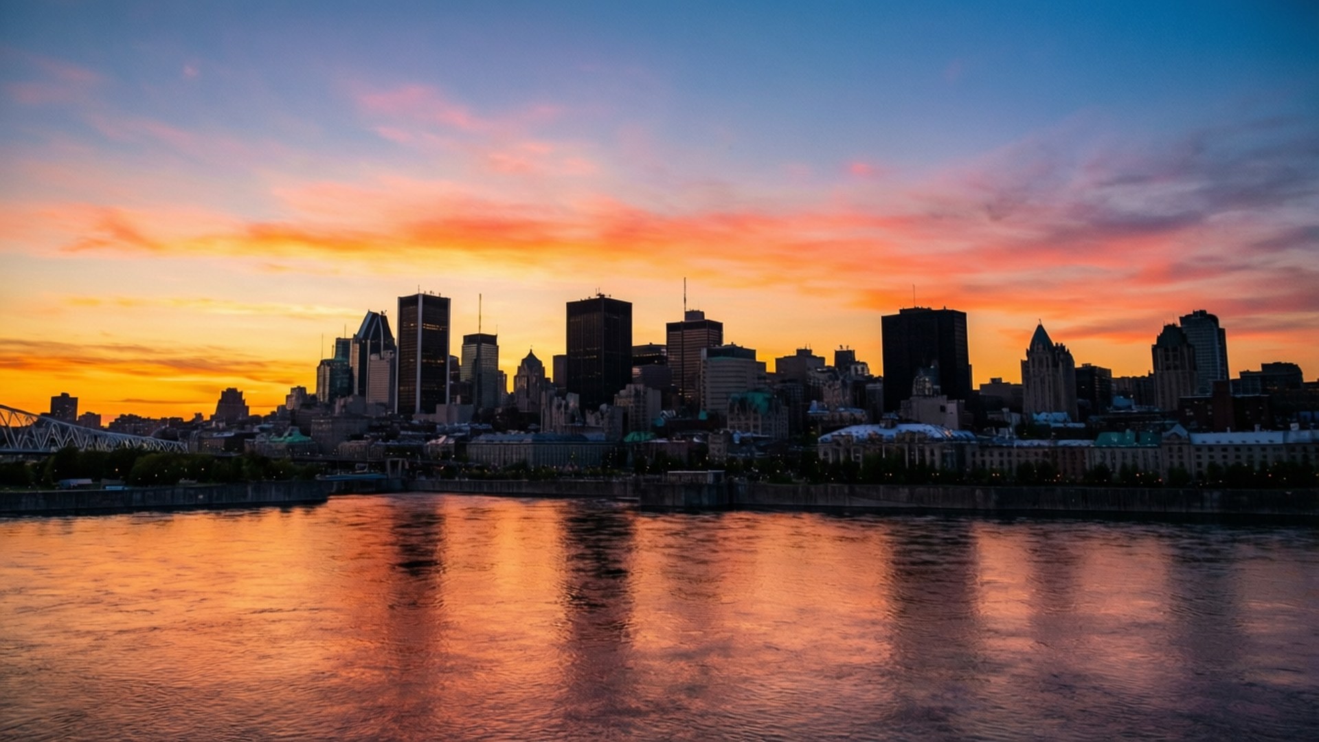 Golden Hour Montreal 壁紙 — 城市天際線在充滿活力的日落襯托下，橙色、粉紅色和藍色的雲彩倒映在平靜的水面上。