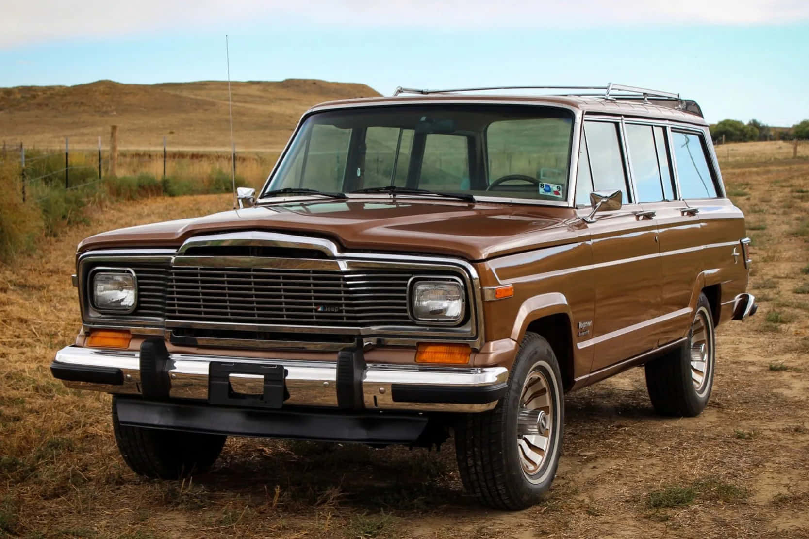 Jeep Wagoneer 桌布 壁紙 — 一輛經典的棕色Jeep Cherokee旅行車停放在乾燥的草地上，背景是晴朗的藍天和起伏的山丘。