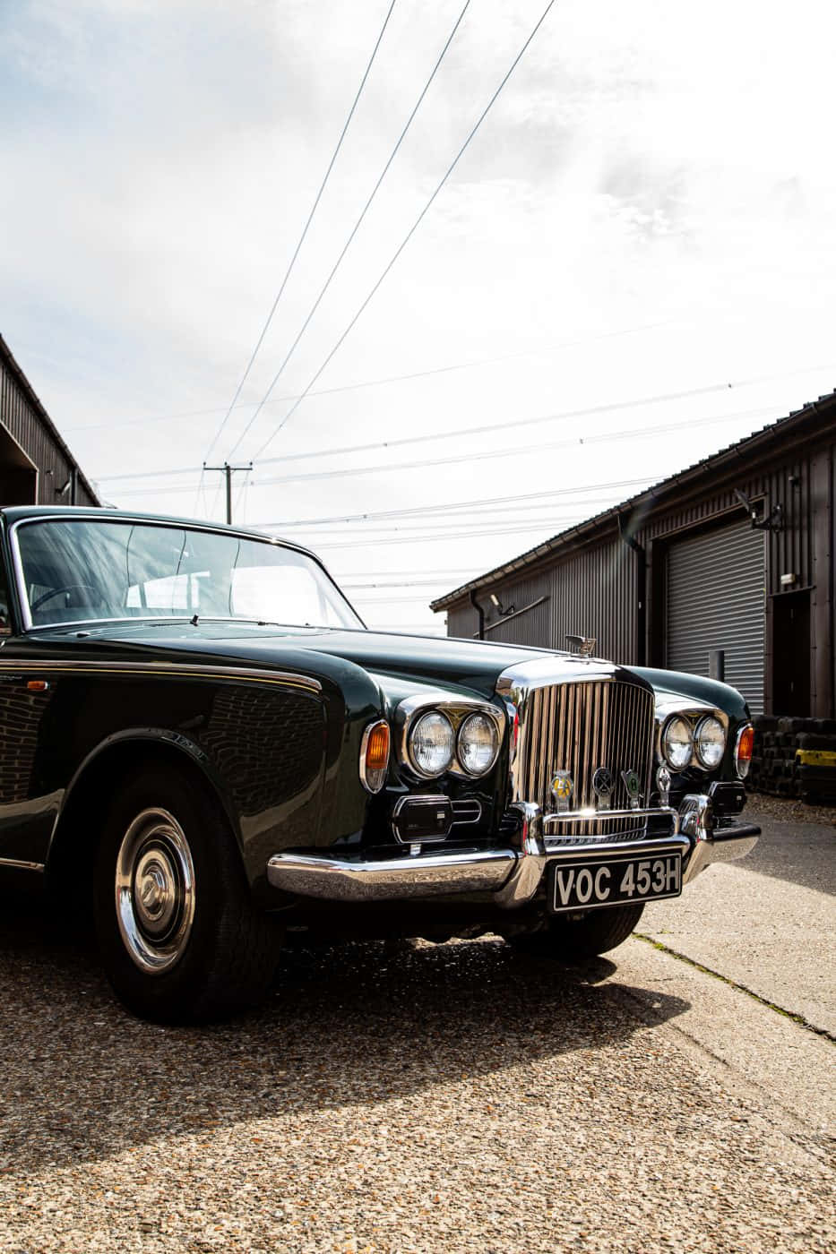 Bentley T1 桌布 壁紙 — 一輛經典綠色賓利，帶有鍍鉻格柵，停泊在工業建築之間的礫石上，天空多雲。