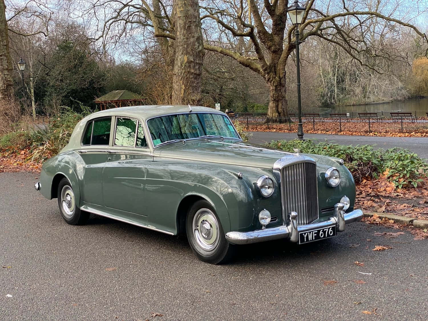 Bentley S2 桌布 壁紙 — 一輛經典綠色賓利房車停泊在落葉滿佈的小徑上，旁邊是寧靜的公園池塘，上方是光禿的冬日樹木。