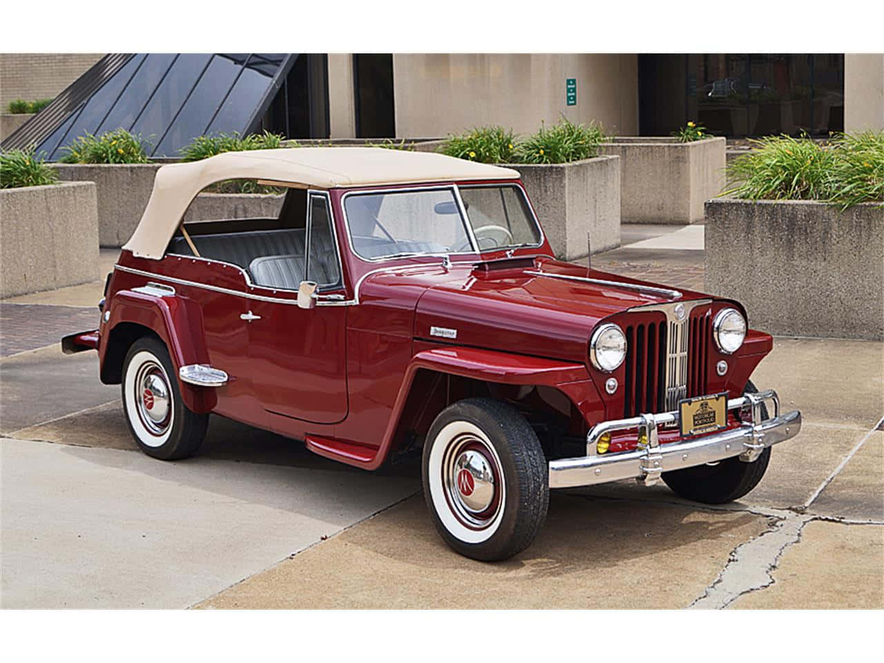 Jeepster 桌布 壁紙 — 經典栗色Jeepster敞篷車配奶油色車頂，停放在現代花盆環繞的混凝土廣場上。