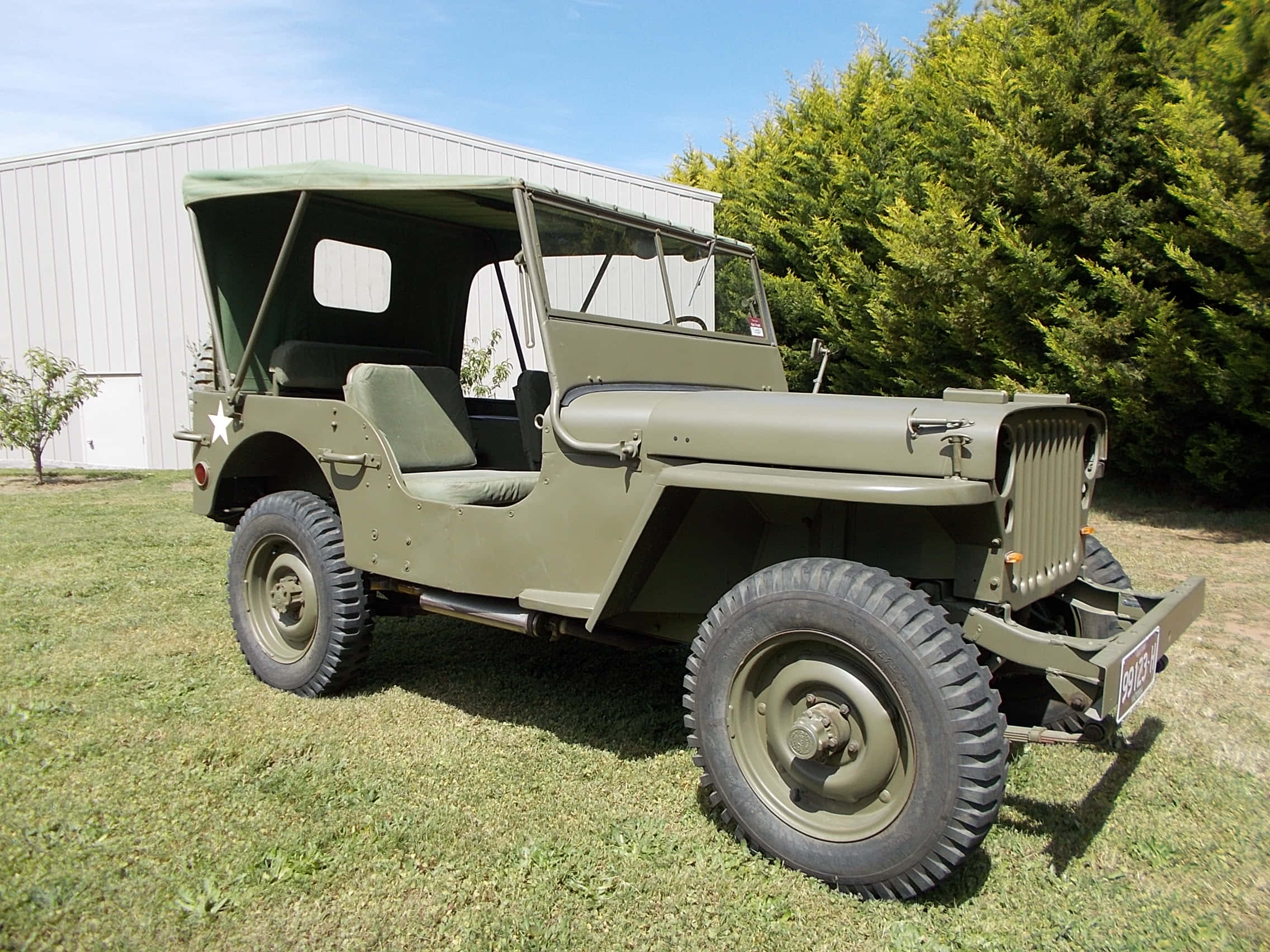 Jeep Willys 桌布 壁紙 — 一輛經典的橄欖綠色軍用吉普車停在草地上，旁邊是白色建築物和茂密的綠樹，天空清澈湛藍。