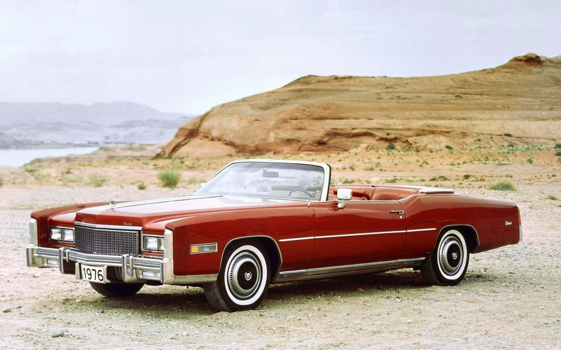 Cadillac Fleetwood 桌布 壁紙 — 一輛經典的紅色1976年凱迪拉克敞篷車停泊在碎石沙漠道路上，背景有岩石山丘和遠處的水面。
