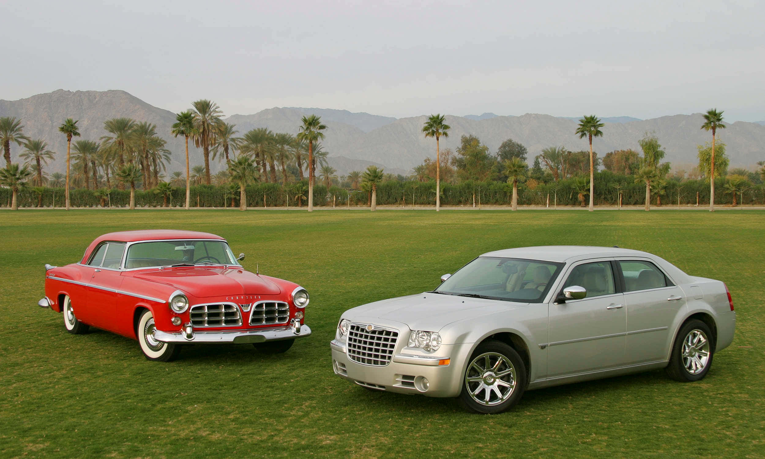 Chrysler 300 桌布 壁紙 — 一輛經典紅色汽車和一輛現代銀色轎車並排停放在茂密的綠色草坪上，背景有棕櫚樹和山脈。