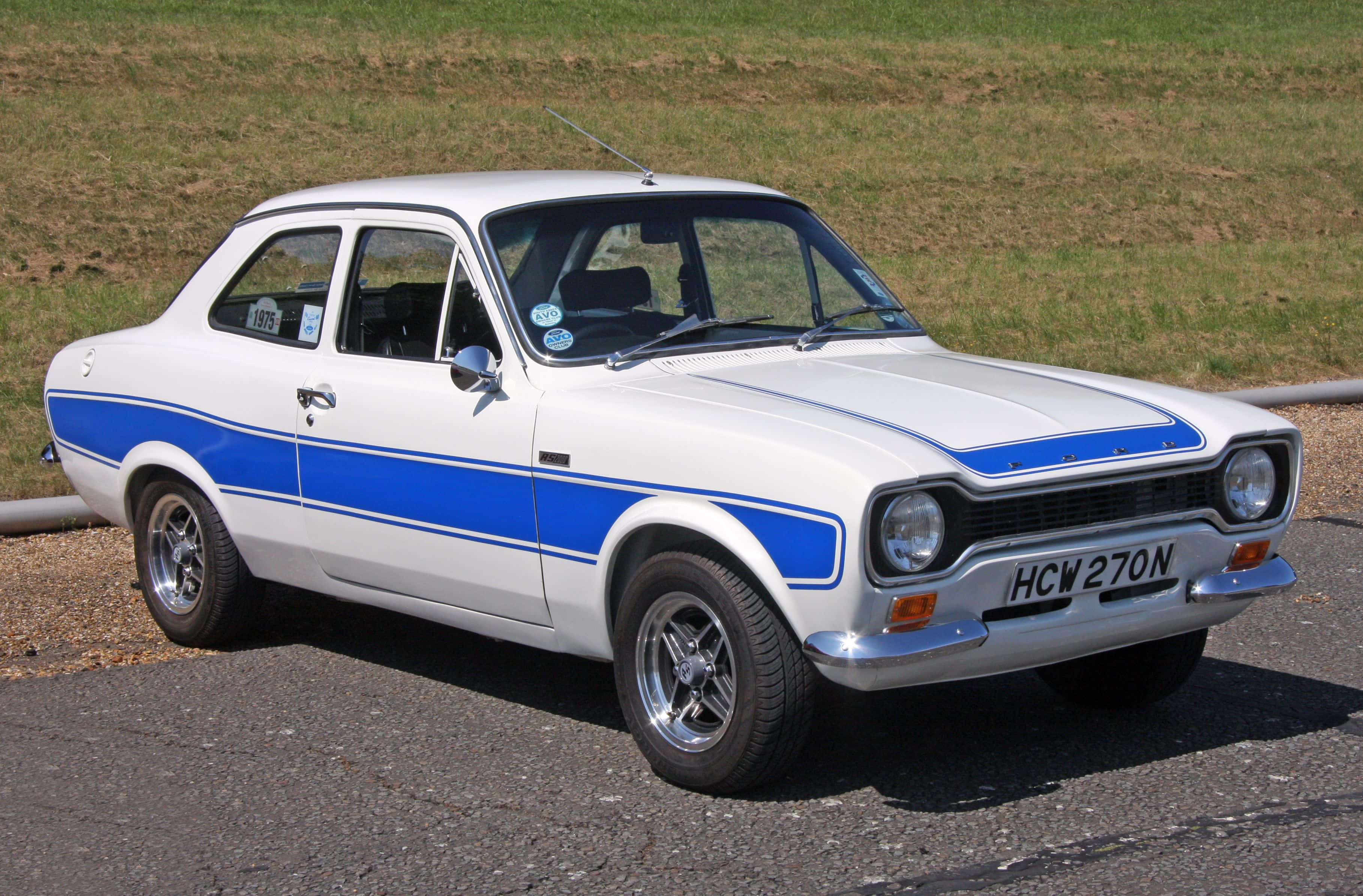 Ford Escort 桌布 壁紙 — 經典白色福特 Capri 帶有醒目藍色賽車條紋，停在陽光普照的草地旁碎石地上。